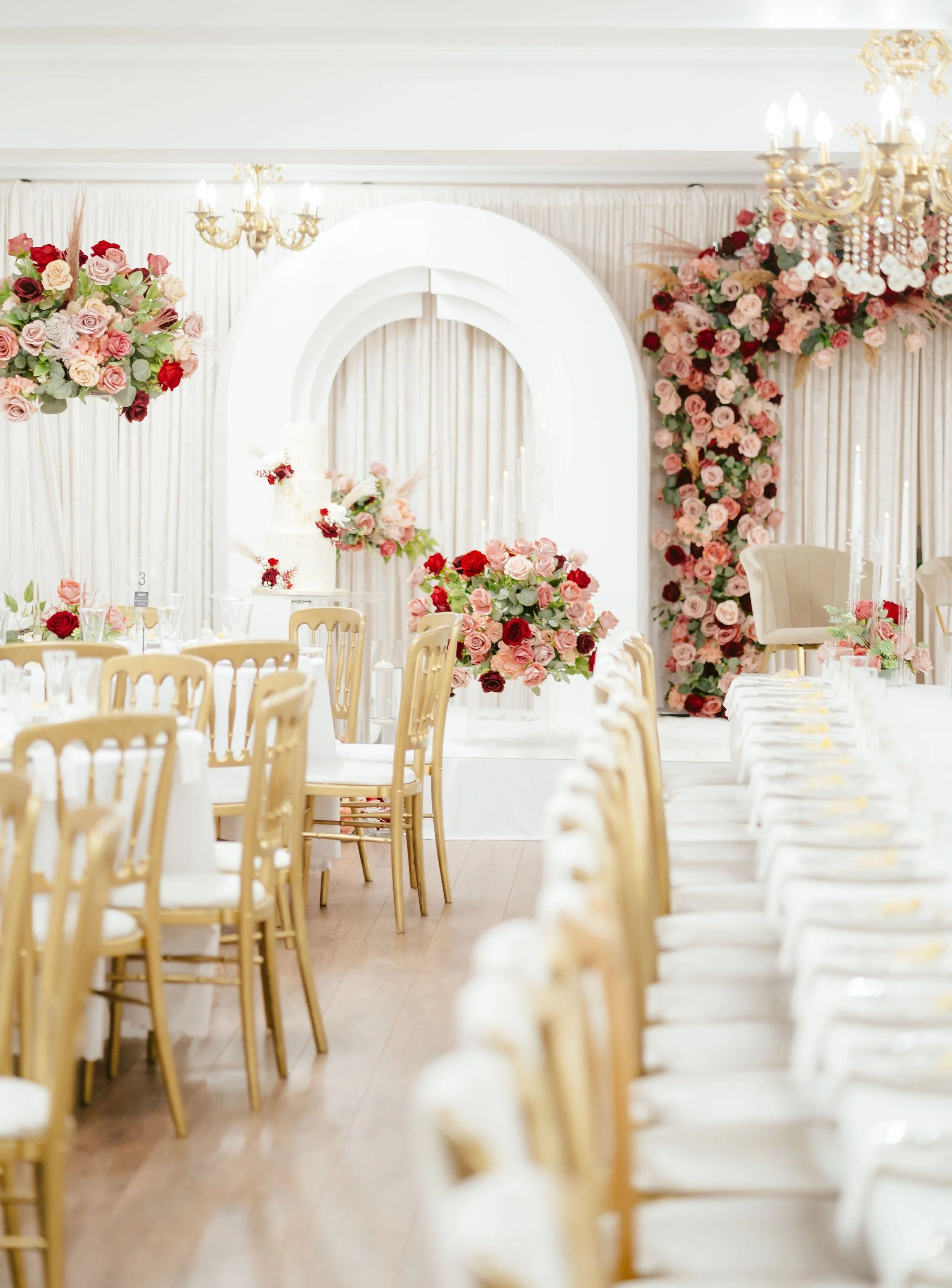 Elegant wedding reception setup with shaded tables and cream-colored chairs, decorated with pink and red roses, amid floral arrangements and a white backdrop with drapes and floral accents.