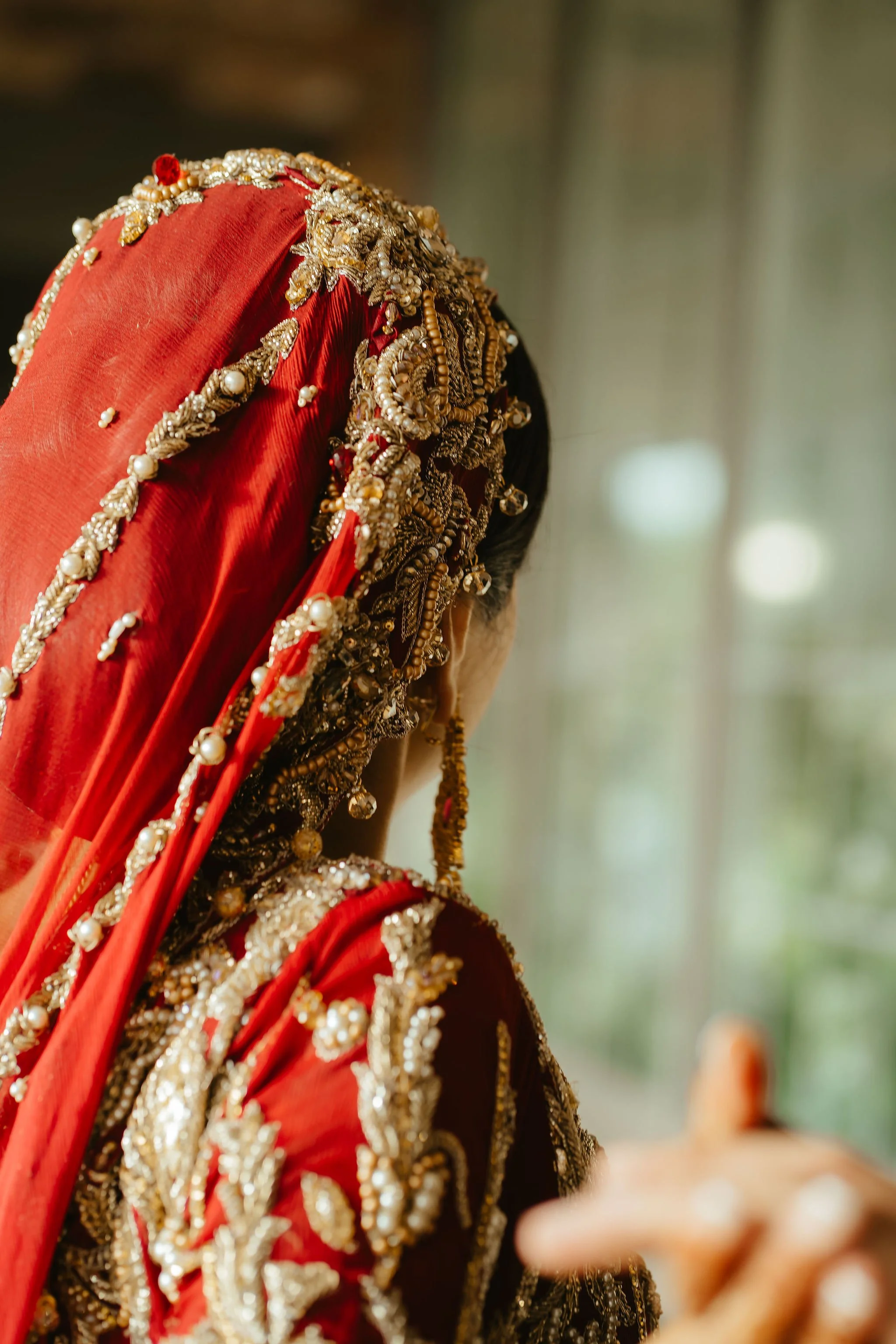 A woman dressed in traditional wedding attire, wearing a heavily embellished red saree with gold and pearl jewelry, seen from the back.