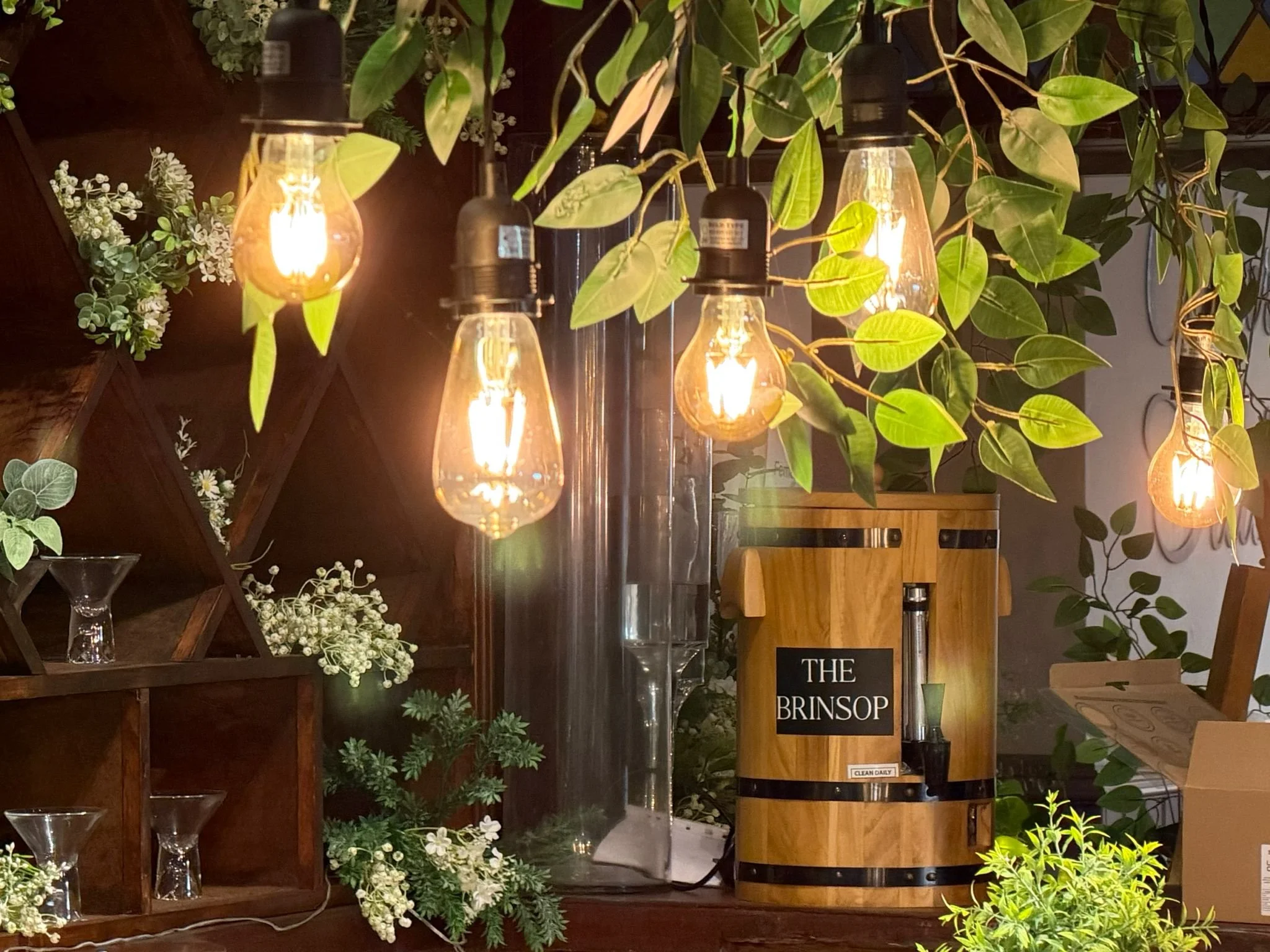 String of hanging Edison-style light bulbs surrounded by green leaves, wooden shelves with small white flowers, and a wooden wine barrel labeled "The Brinsop" in a cozy indoor space.