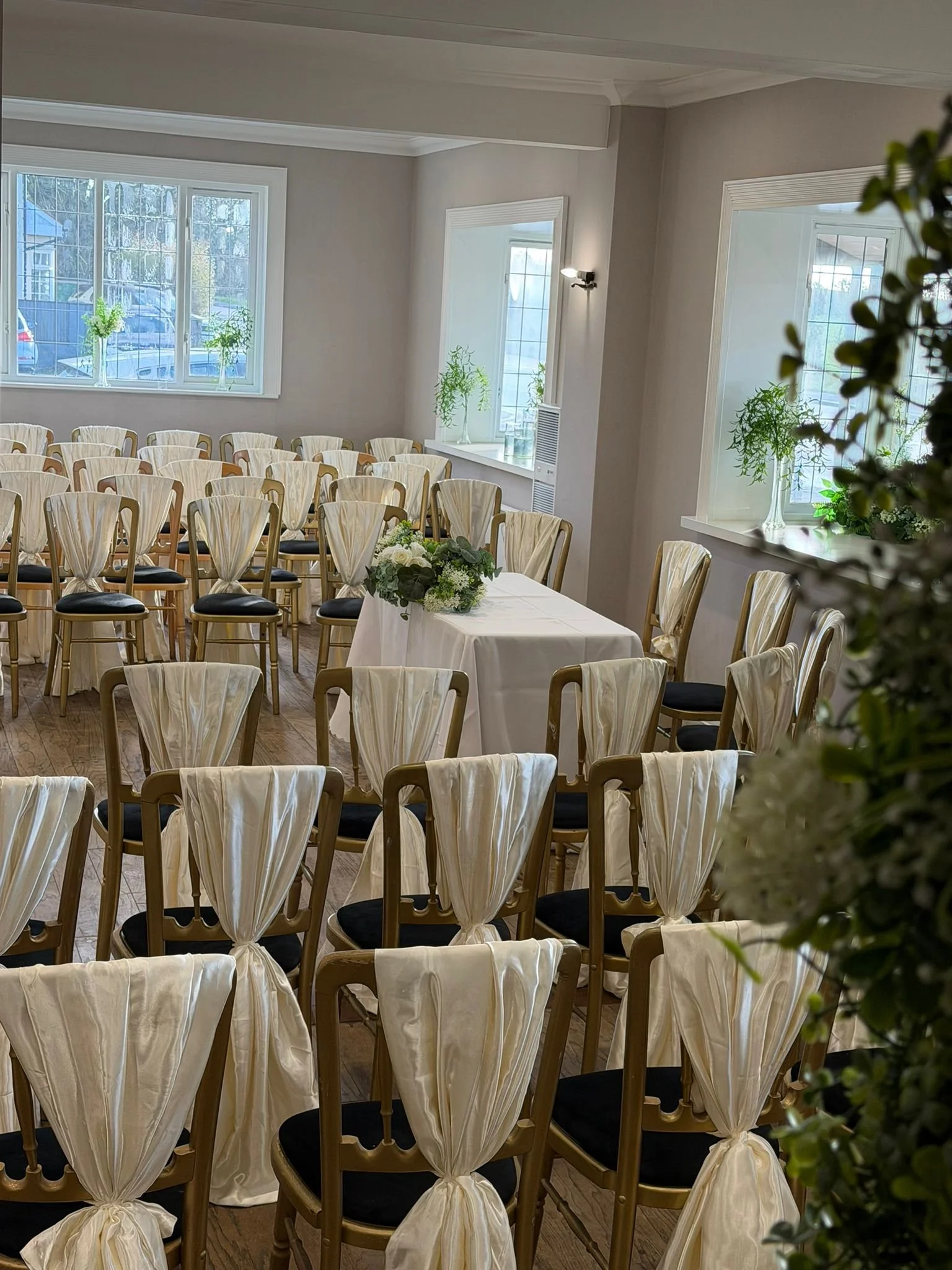 Empty wedding ceremony room with gold chairs, decorated with cream-colored sashes, arranged in rows with an aisle in the center. A table with floral arrangement is at the front. White walls, large windows with potted plants, and hardwood floors.