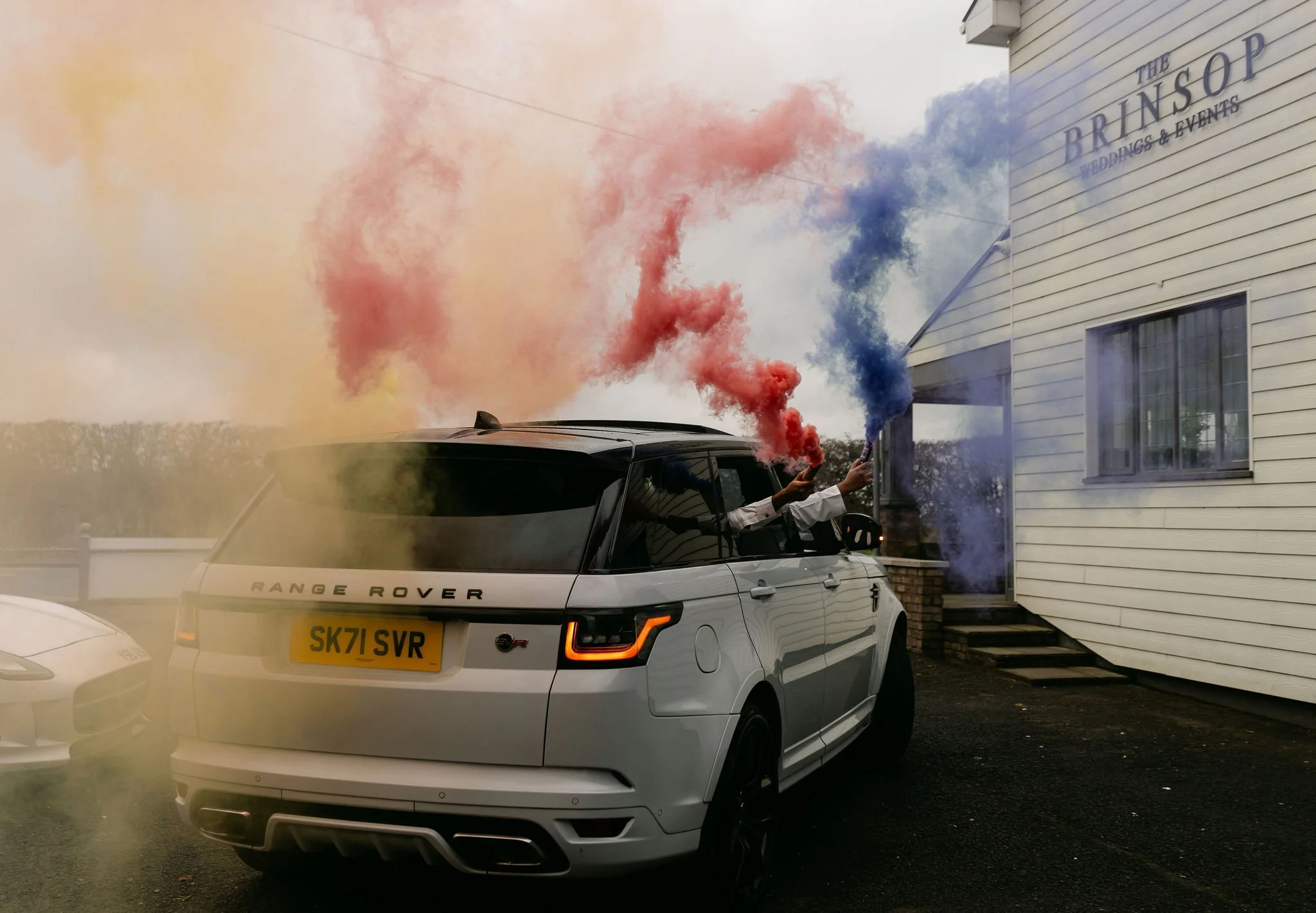 White Range Rover SUV with colorful smoke coming from people inside. The smoke is red, pink, yellow, and blue. The SUV is parked outside a building with a sign that reads "The BRINSOP wedding & events." There is another car partially visible on the l