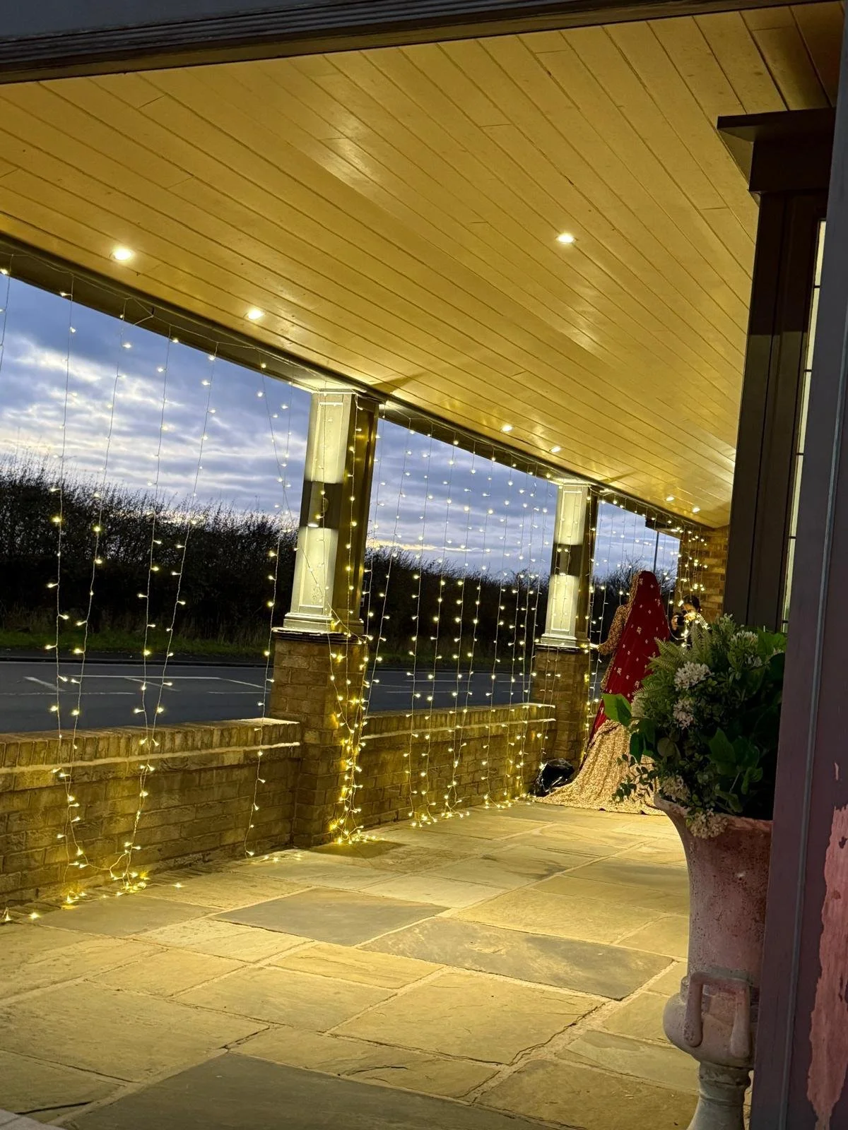 A porch decorated with string lights, a potted plant with greenery and white flowers, and a person dressed in red traditional clothing with gold embellishments, facing away towards the scenery outside on a cloudy evening.