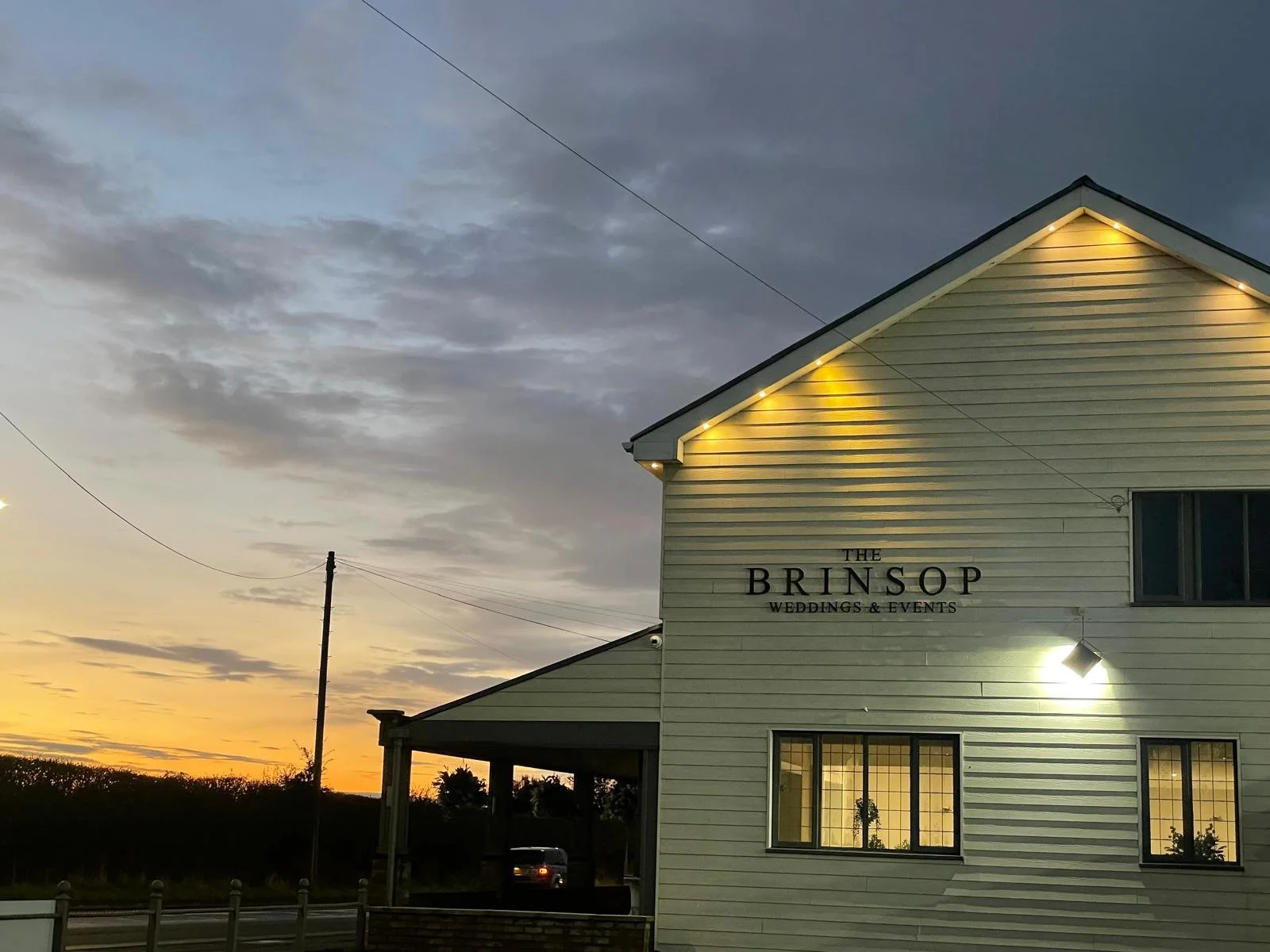 A two-story white building with a sign that reads "The Brinsop Weddings & Events" at sunset, with lit exterior lights and a car parked underneath a porch, against a backdrop of a cloudy sky and a horizon with trees.