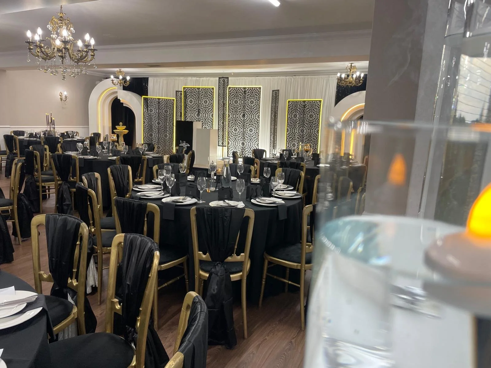 Elegant banquet hall set up with round tables covered in black tablecloths, gold and black chairs, and table settings including plates, glasses, and silverware. Chandeliers hang from the ceiling, and decorative partitions with intricate patterns are 