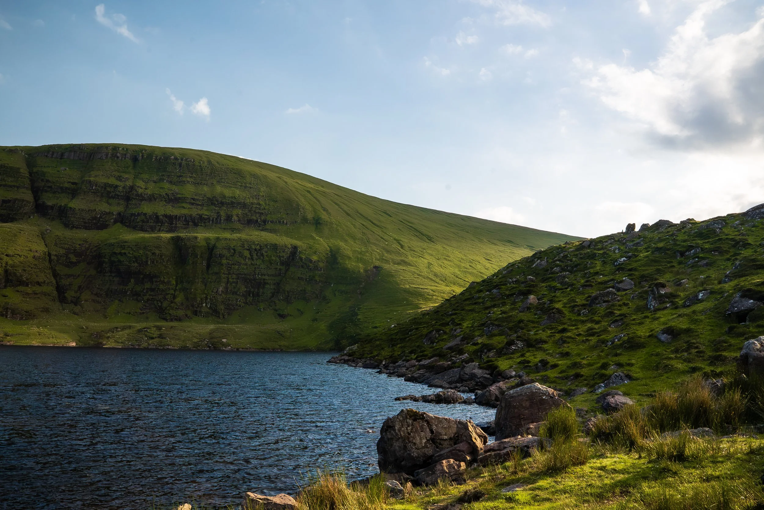 A lake surrounded by green hills and rocks, with a partly cloudy sky.