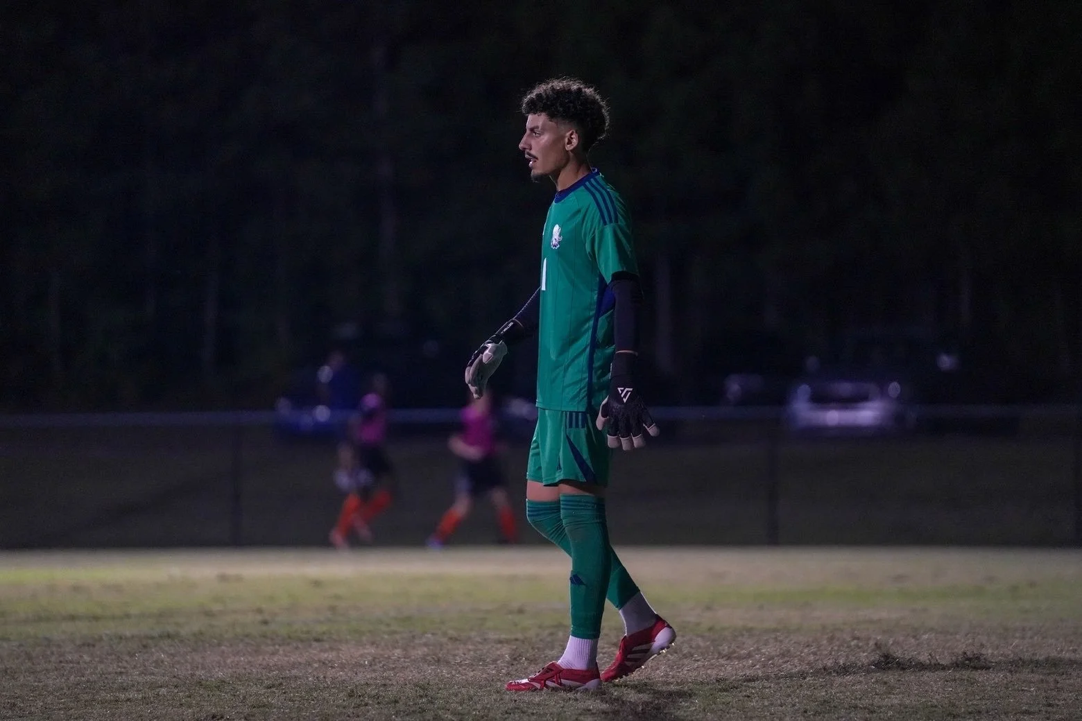 A soccer goalkeeper standing on the field at night, wearing a green uniform with goalkeeping gloves, looking focused.