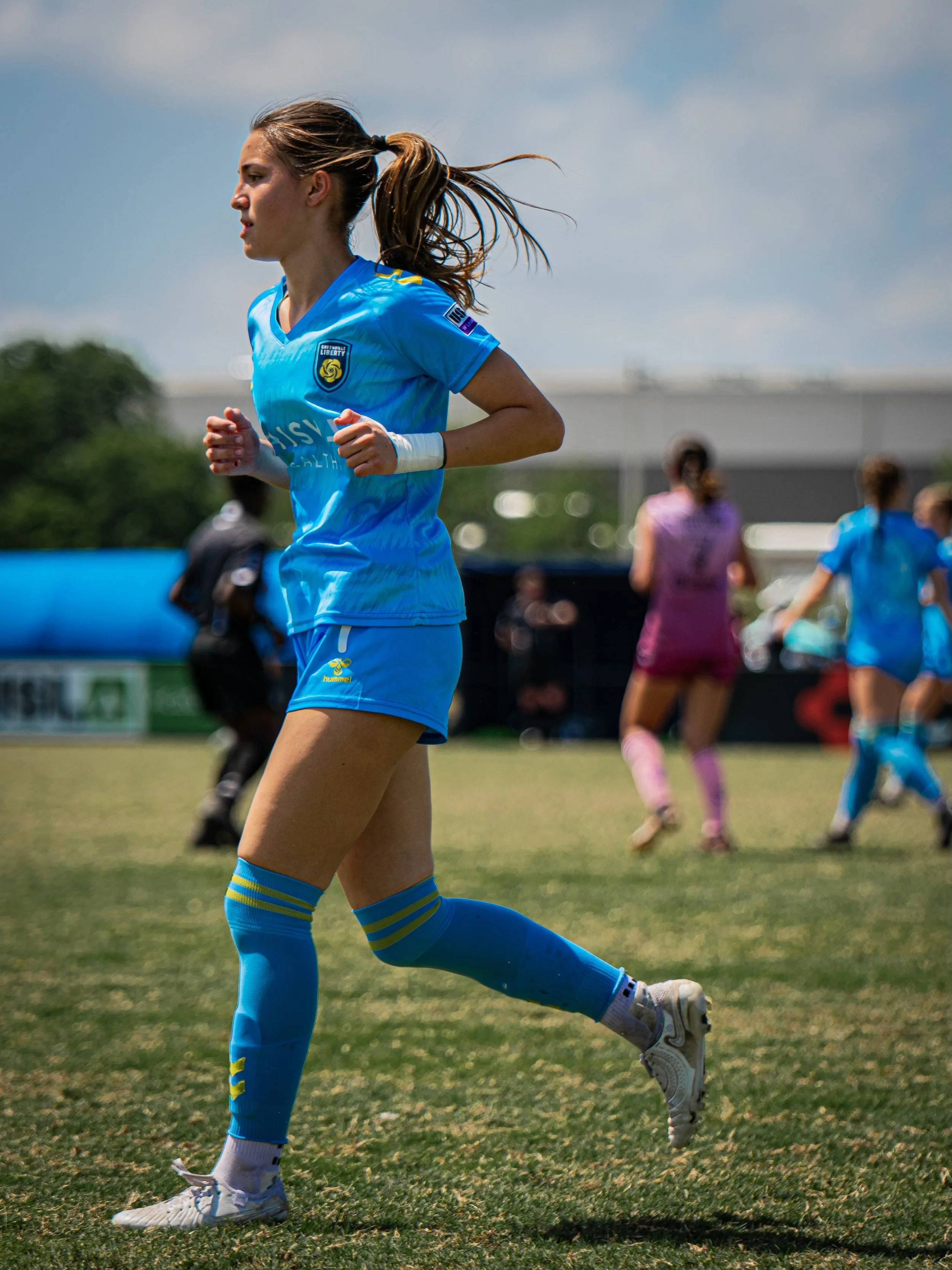 A female soccer player in a blue uniform running on the field during a game, with other players and a referee in the background.