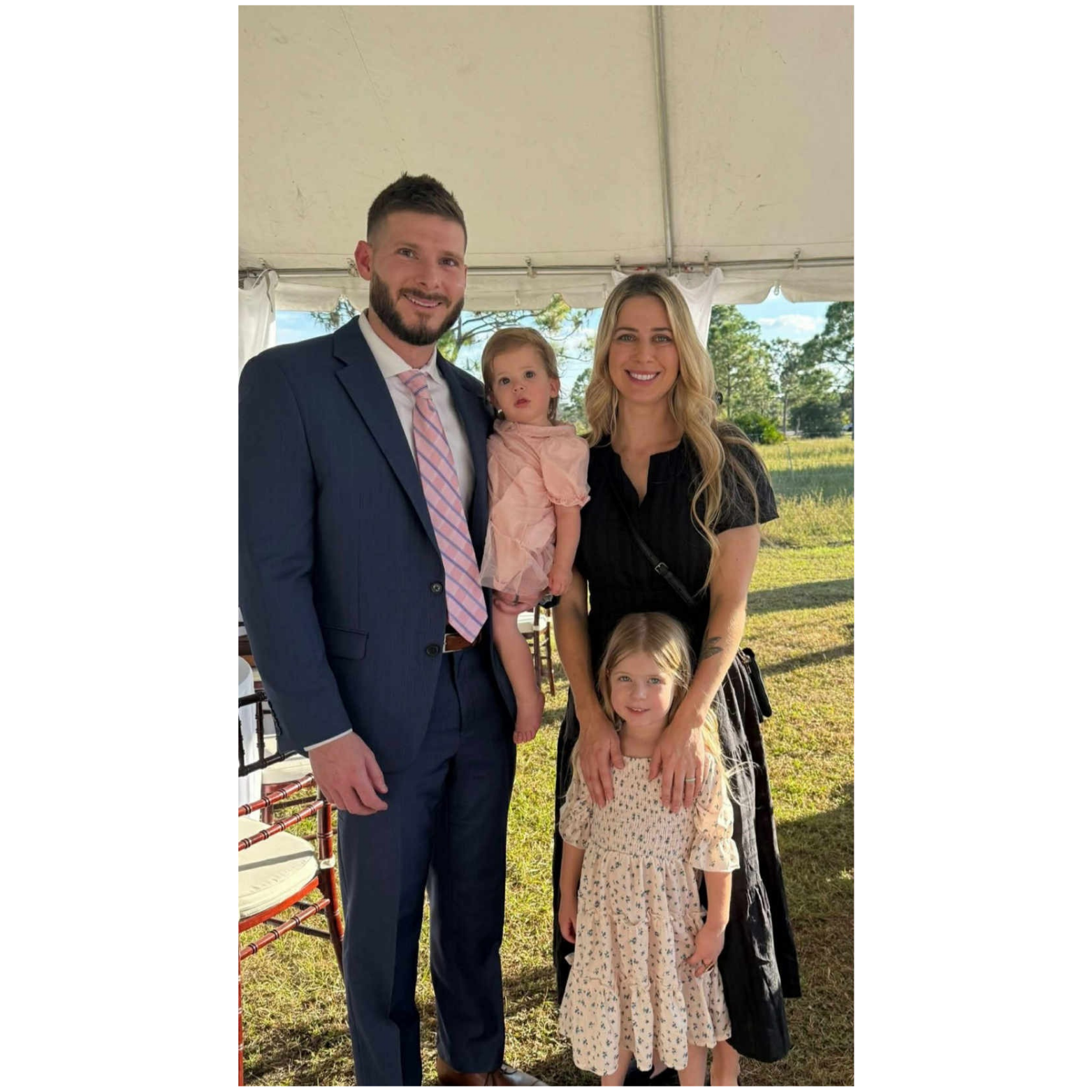 A family of four posing outdoors under a tent on a sunny day. The man is wearing a dark blue suit with a pink and white striped tie. The woman is in a black dress. The little girl in front is wearing a cream-colored dress with a floral pattern. The toddler girl is dressed in a pink dress and is being held by the man.
