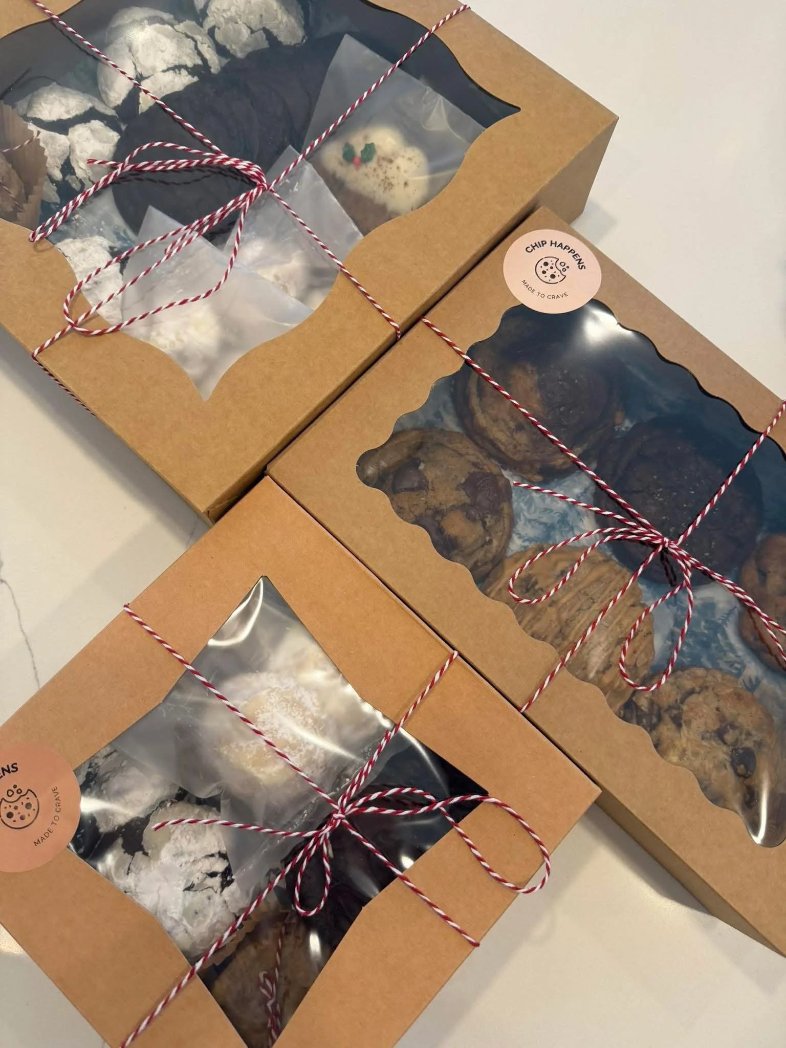 Three brown bakery boxes with clear windows showing cookies inside, secured with red and white twine, on a white surface.