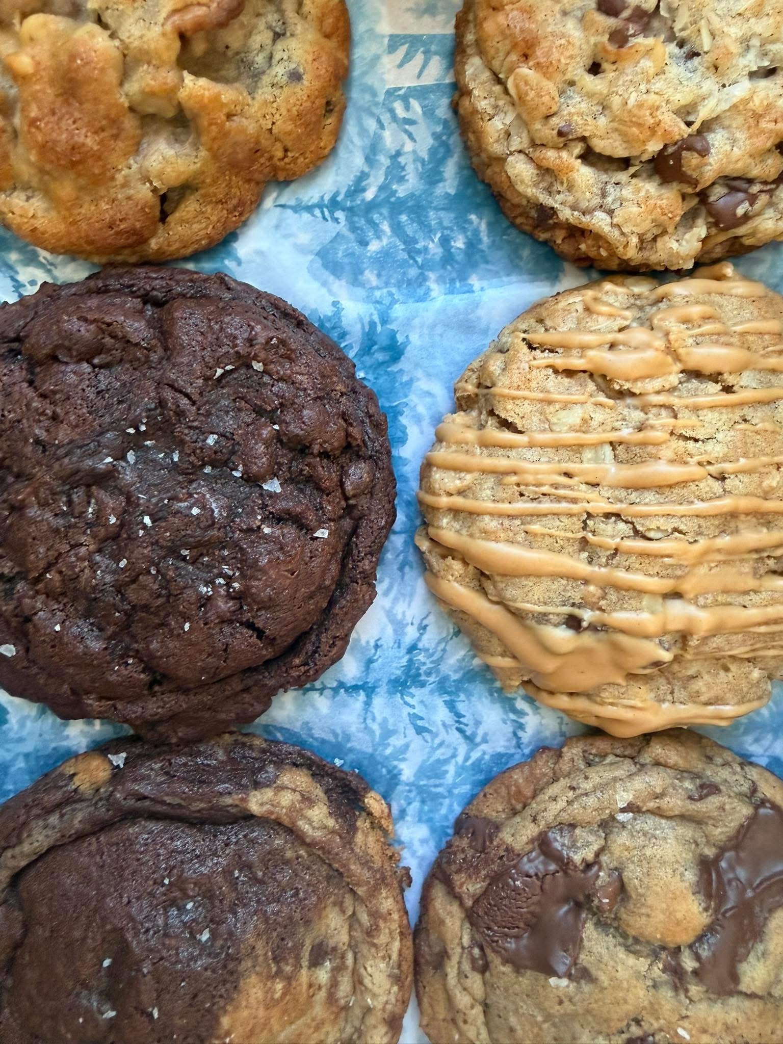 Assorted cookies on a blue and white patterned paper, including chocolate chip, oatmeal, peanut butter, and double chocolate cookies.