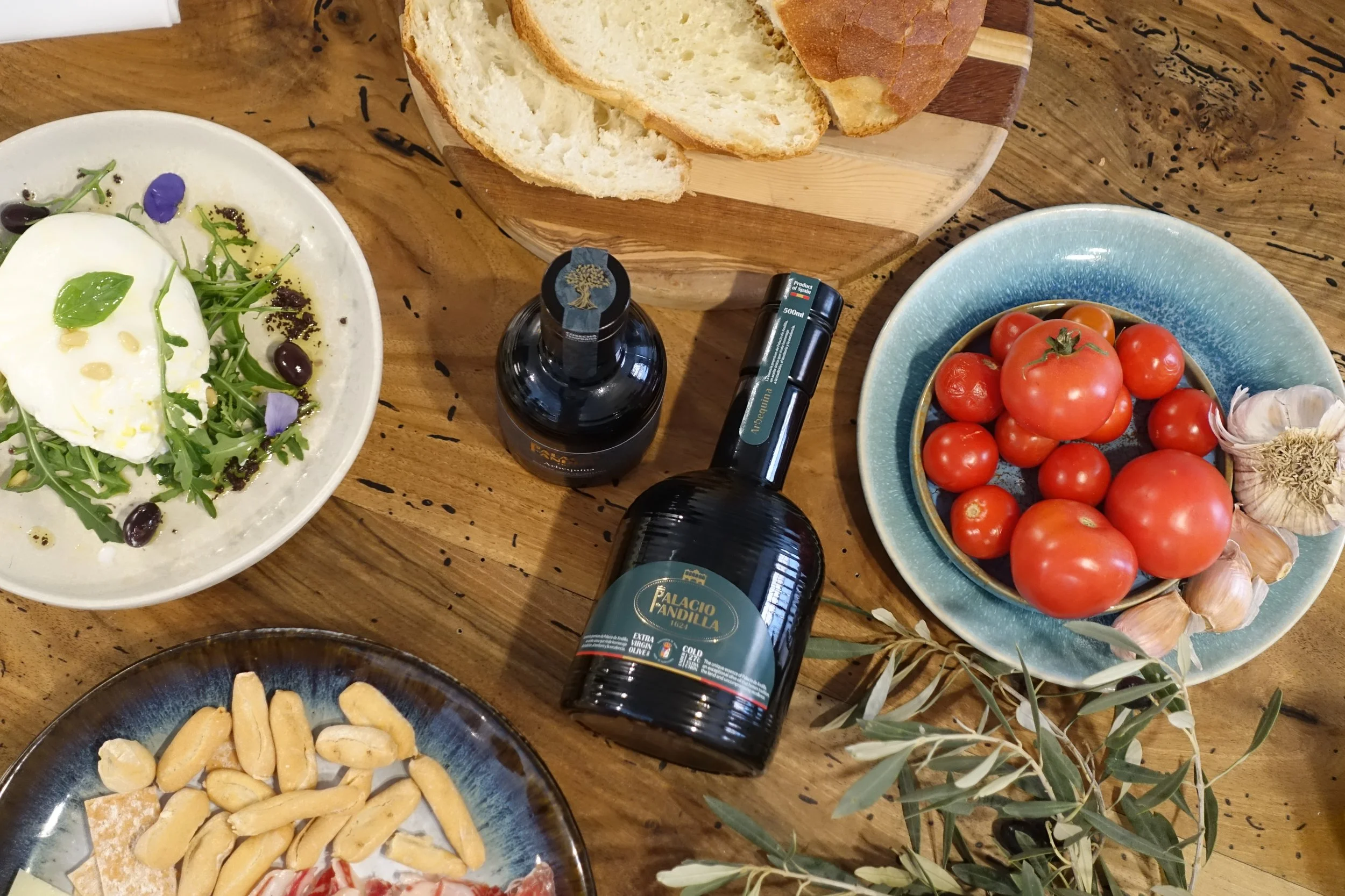 Vista superior de una mesa con platos de ensalada, pan, tomate, ajo, botella de vino y aperitivos.