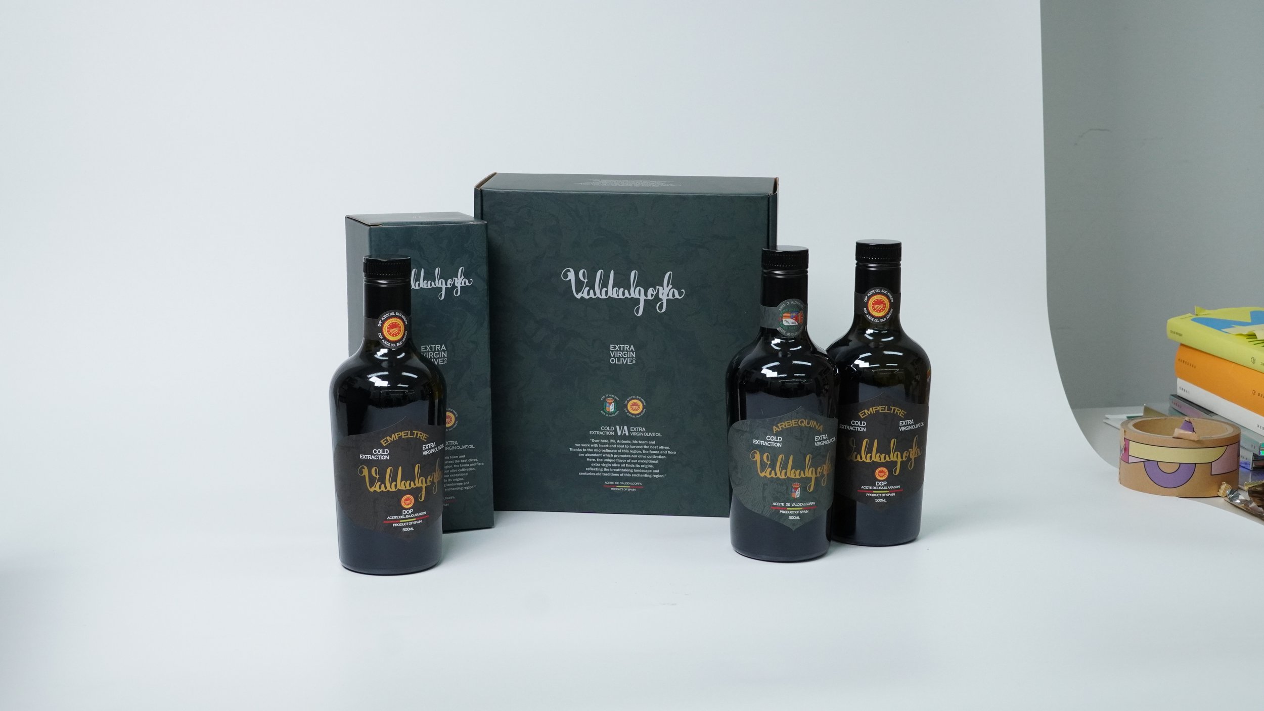 Tres botellas de aceite de oliva extra virgen sobre una mesa blanca, con cajas y libros en el fondo a la derecha.