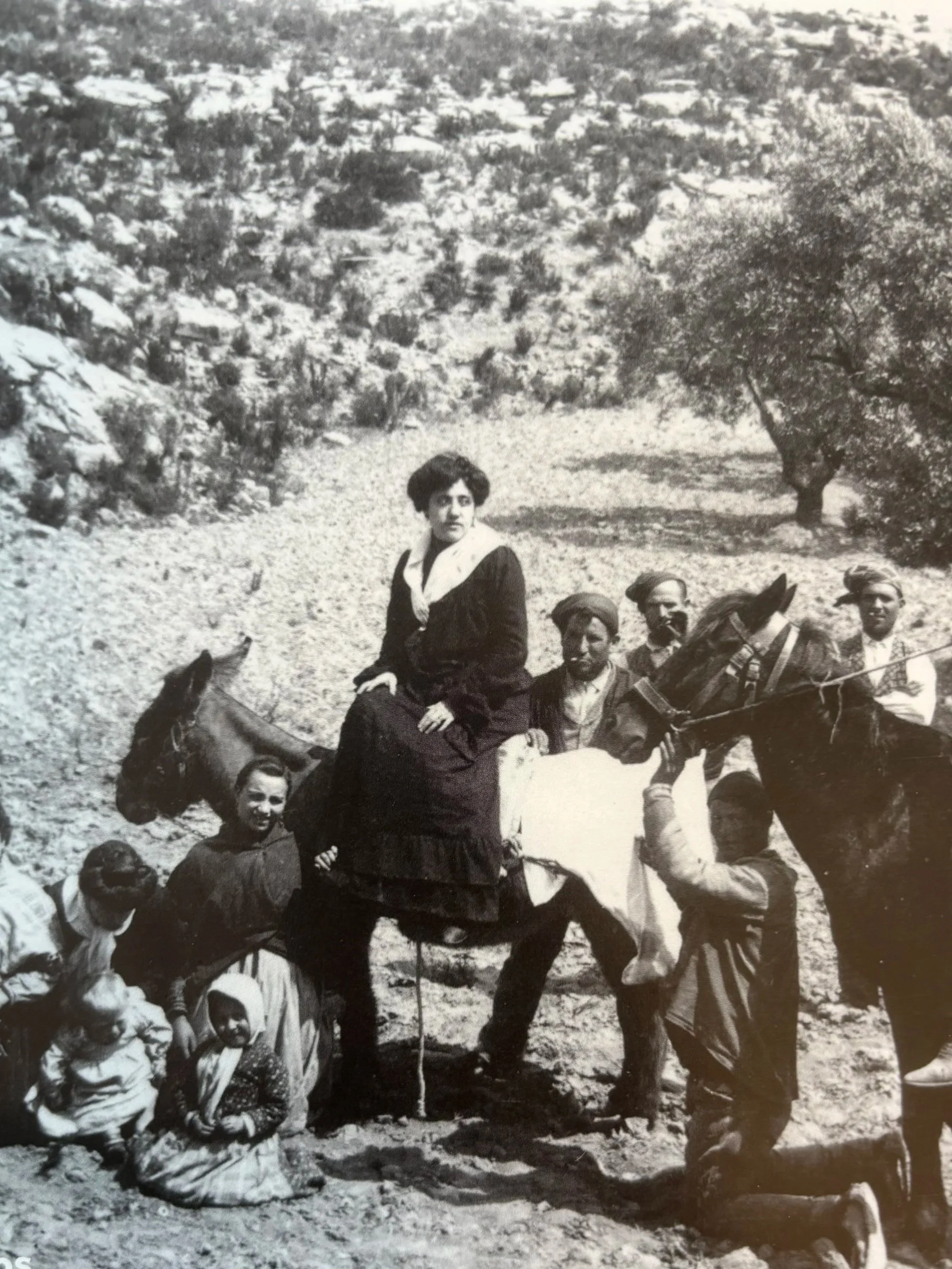 Una mujer montada en un caballo rodeada de varias personas, en un paisaje rural con vegetación y árboles.