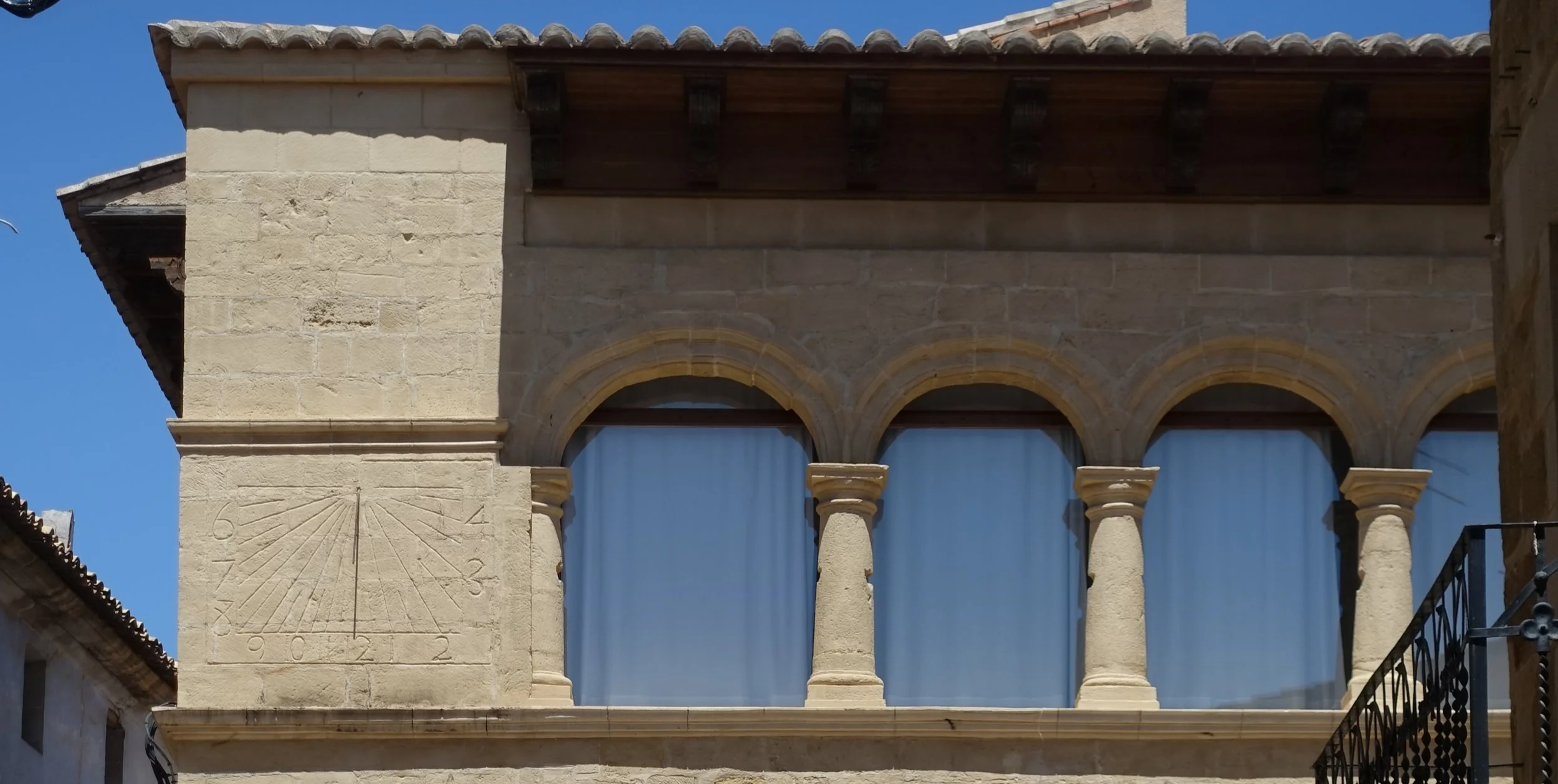 Edificio de piedra con ventanas arqueadas, columnas en el balcón y un reloj de sol en la esquina izquierda
