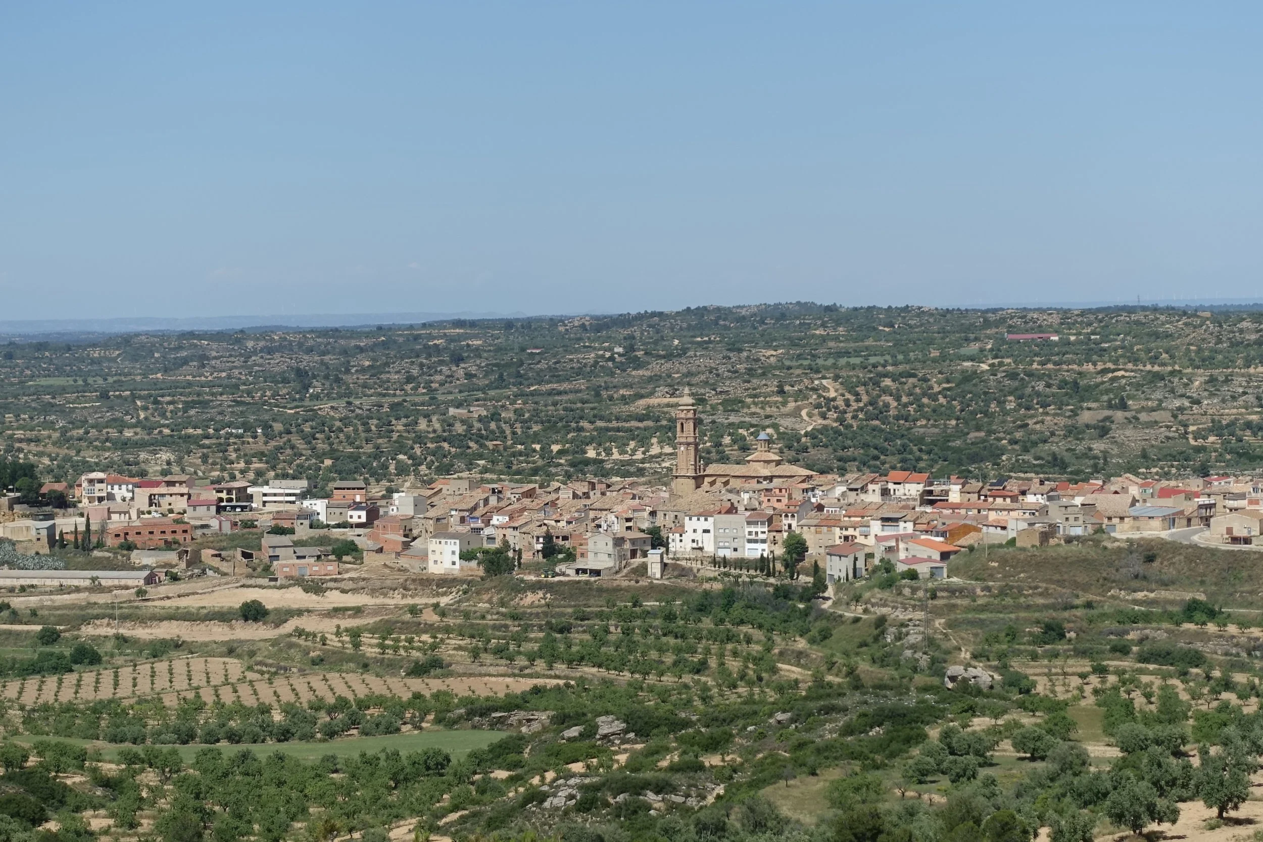 Vista panorámica de un pueblo rodeado de sembradíos y colinas, con una iglesia de torre alta en el centro, bajo un cielo despejado.