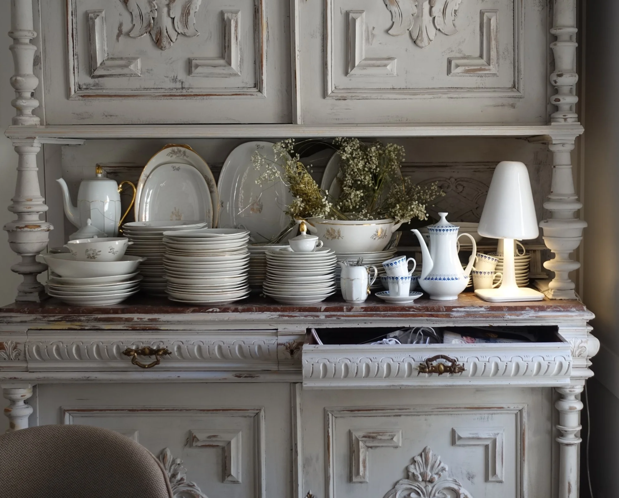 Vajilla blanca decorada con oro y azul en un mueble antiguo, con flores y una lámpara pequeña de mesa.