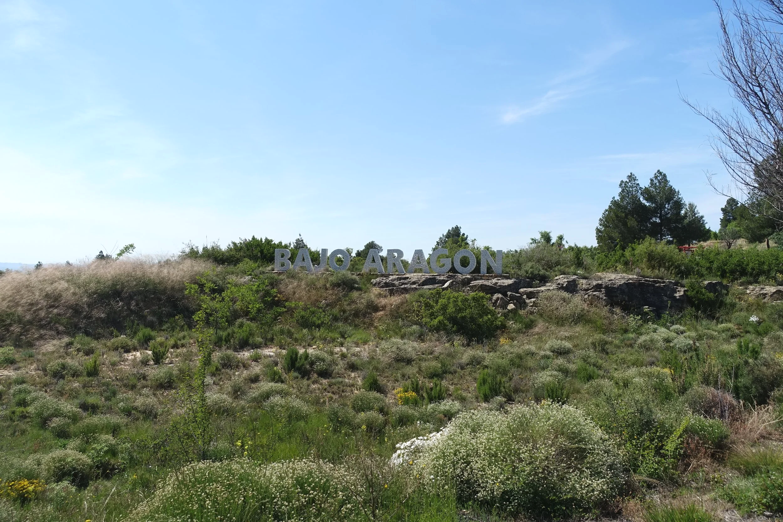 Cartel que dice 'BAJO ARAGÓN' en un paisaje de vegetación y rocas, con árboles y un cielo despejado.