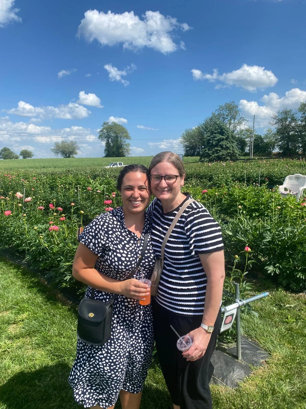 Two women smiling and holding drinks in front of a flower field with pink flowers and a clear blue sky with scattered clouds.