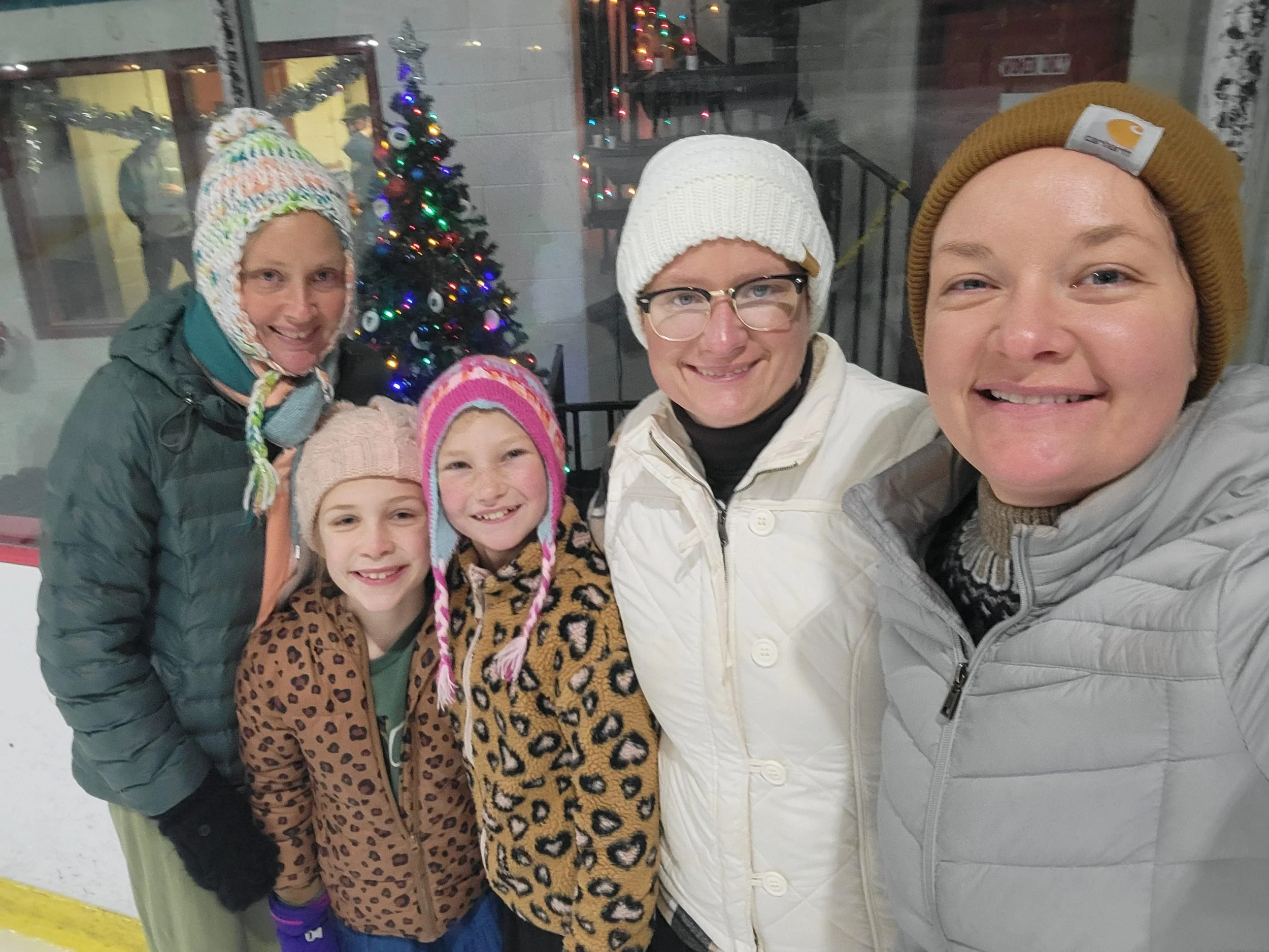 Group of five women and two girls taking a selfie together in front of a decorated Christmas tree at an ice skating rink, all wearing winter coats and hats.