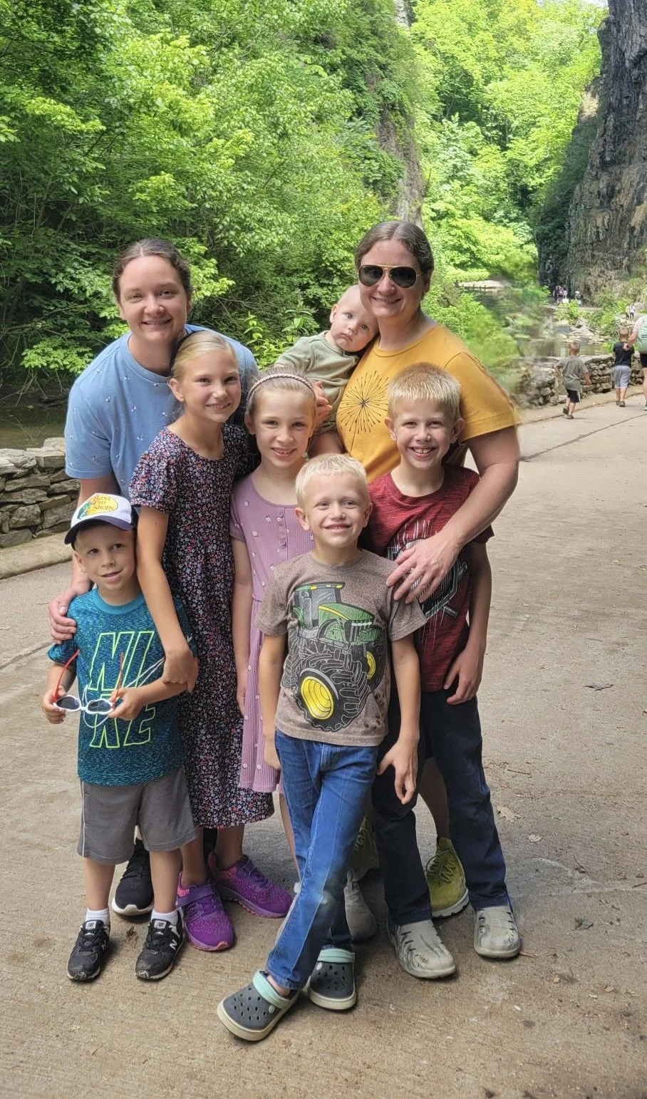 A group of nine people, including adults and children, posing outdoors in front of a lush green forest with large rock formations.