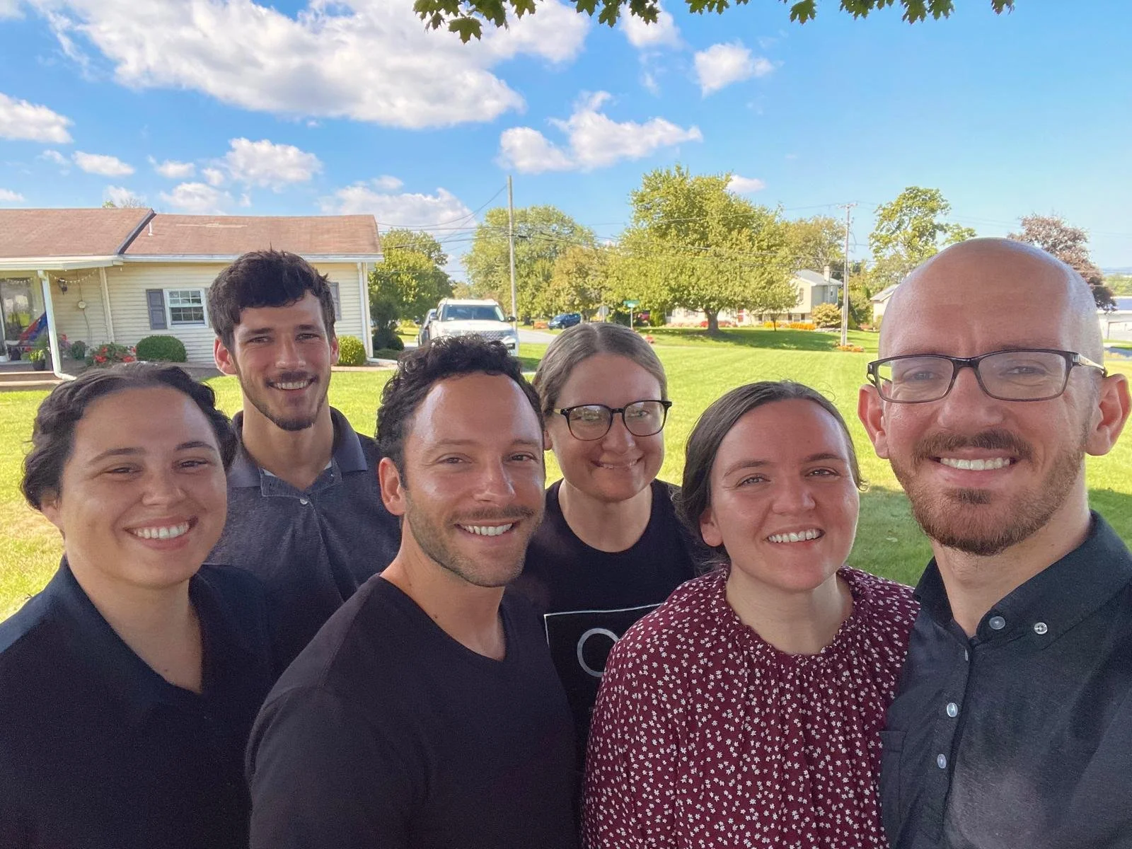 Group of six smiling people taking a selfie outdoors on a sunny day with a house, trees, and a grassy yard in the background.