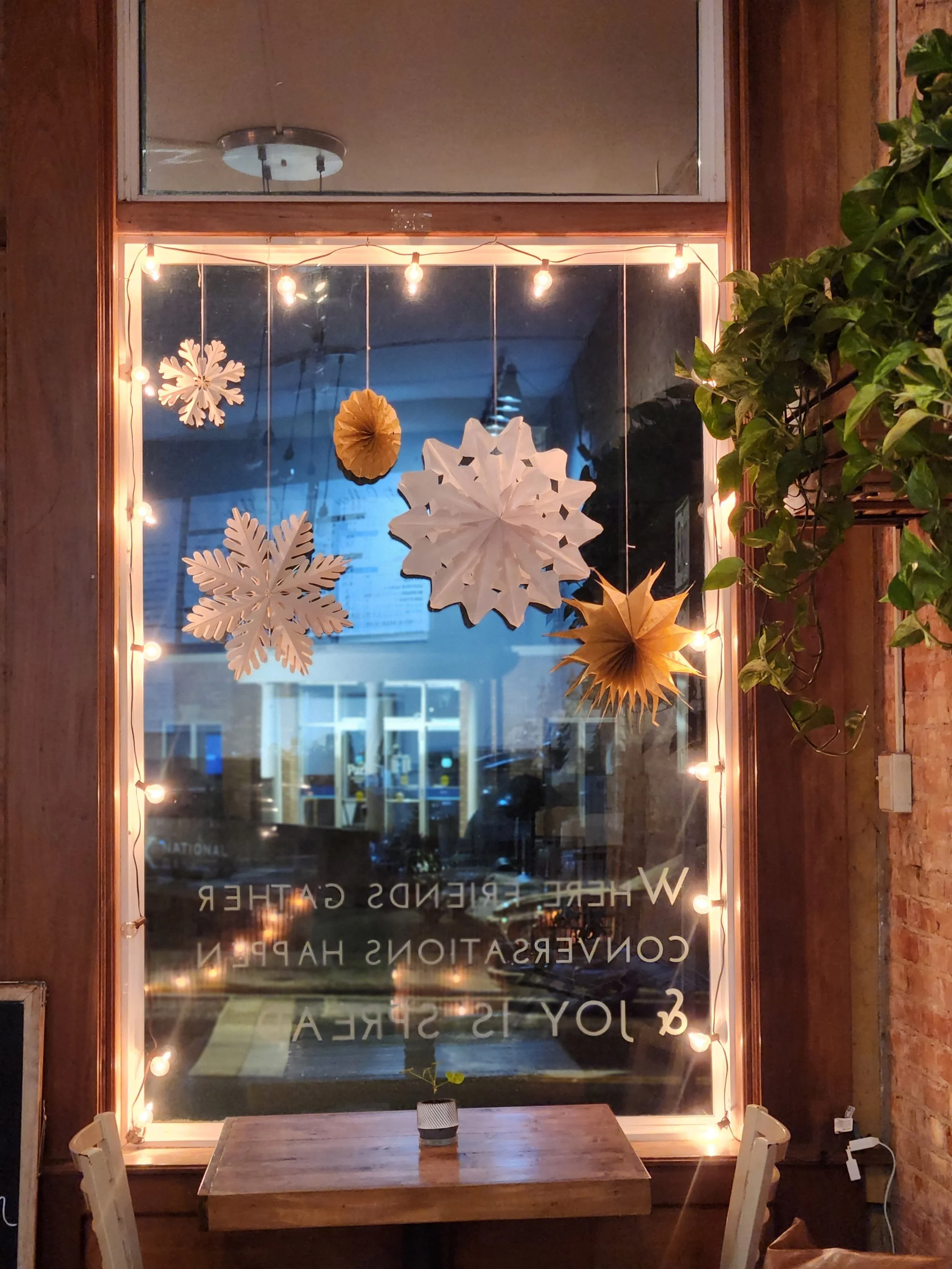 Decorative window display with hanging paper snowflakes and ornaments, string lights, a small wood table with a potted plant, and a brick wall on the side.