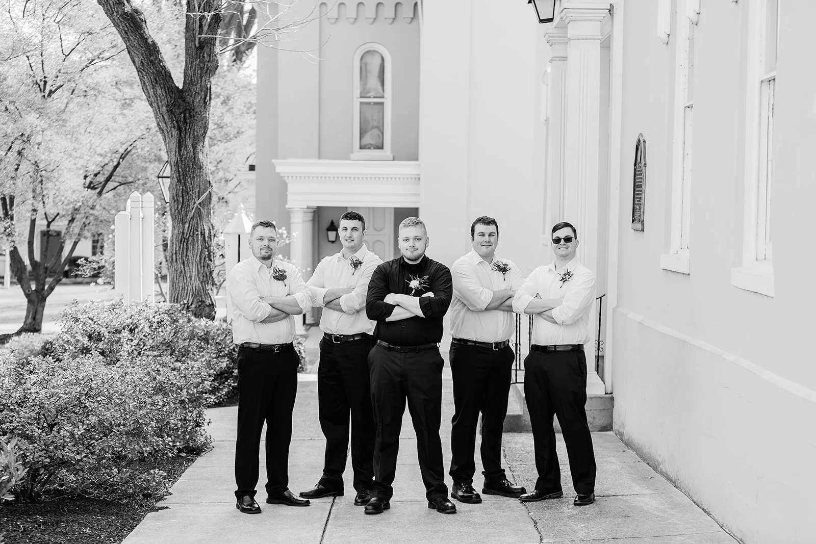 A group of five men standing outdoors on a sidewalk in front of a building. They are dressed in formal shirts with boutonnières, with the man in the center wearing a black shirt. All have their arms crossed and are smiling.