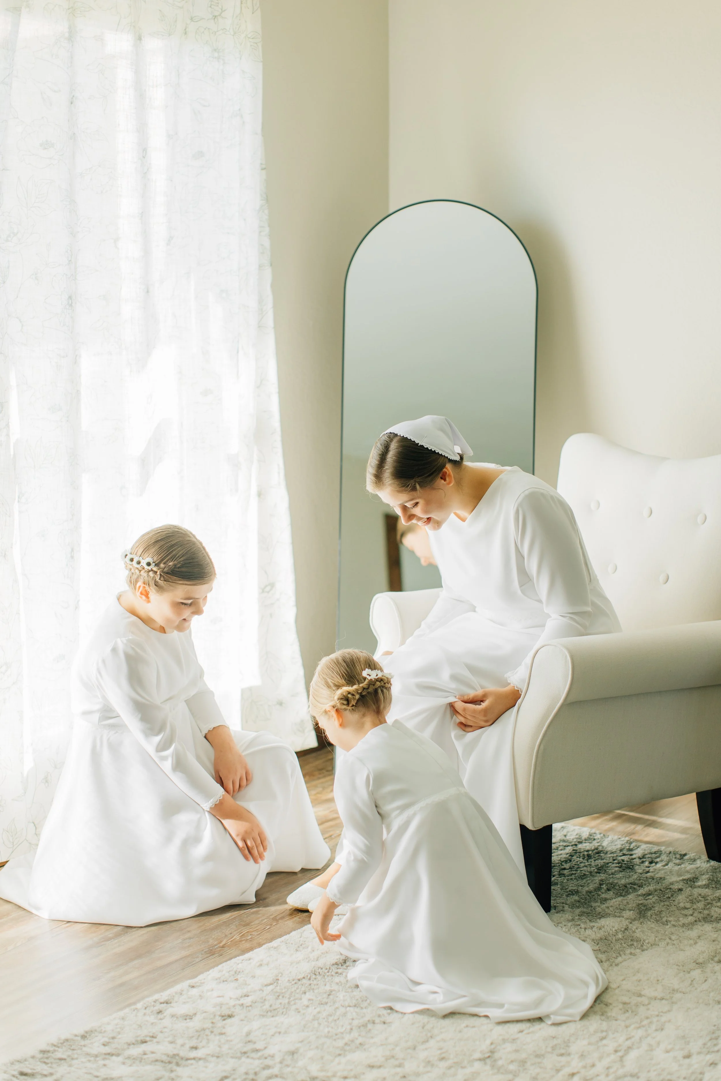 A woman and three young girls dressed in white, possibly preparing for a special occasion, in a bright room with a mirror, a white armchair, curtains, and a rug.