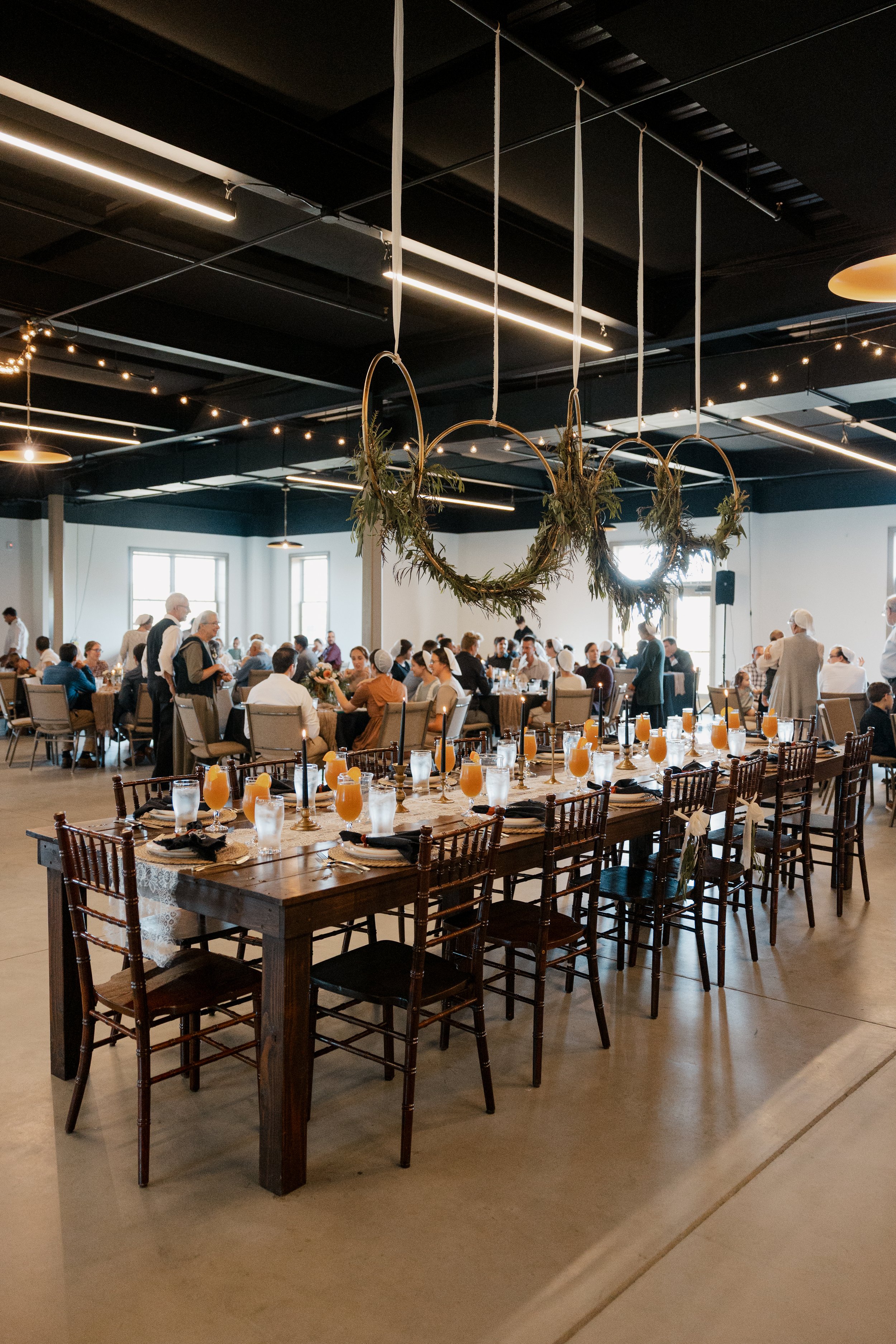 Wedding reception with decorated tables, hanging greenery, and guests seated and mingling indoors.