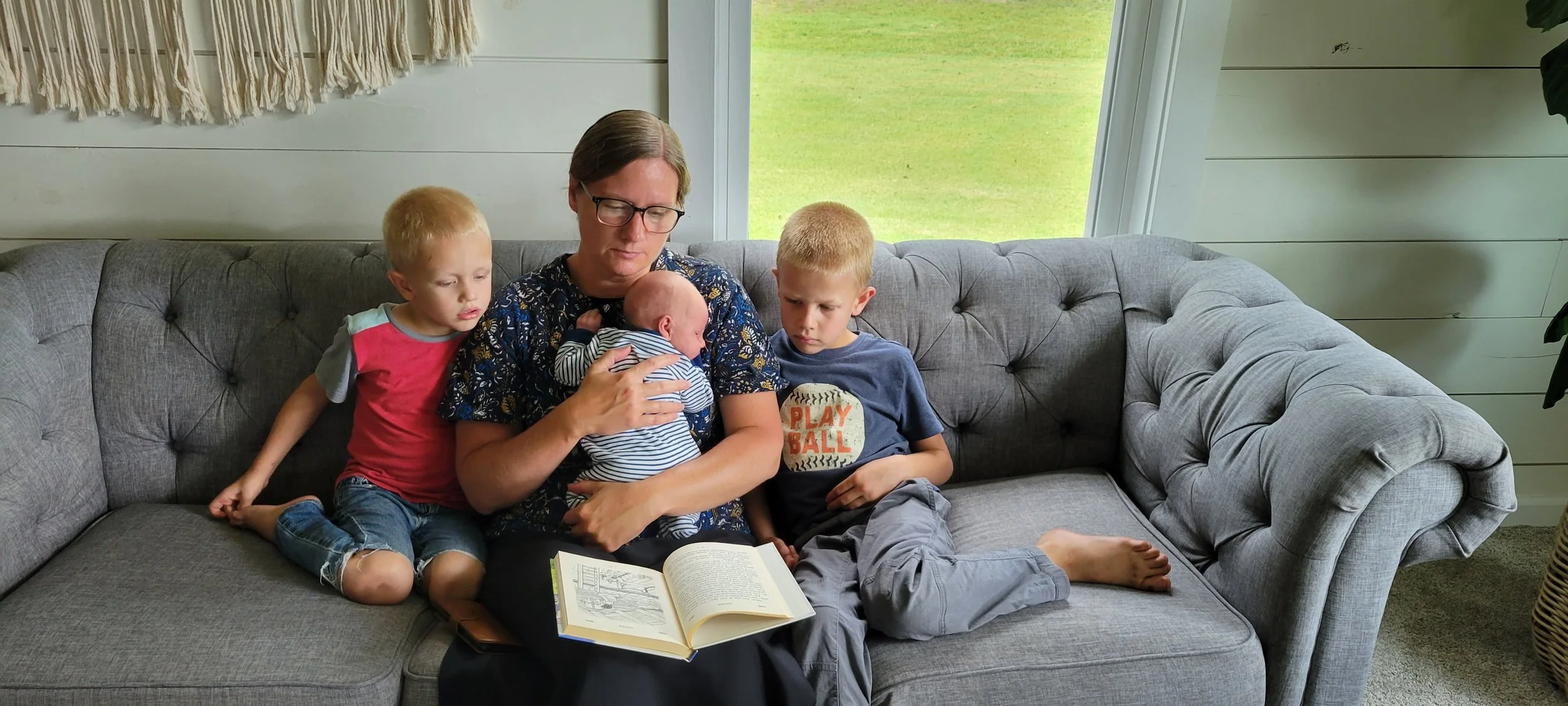 A woman reading a book to three young children on a gray couch in a room with a large window and green outdoor scenery.