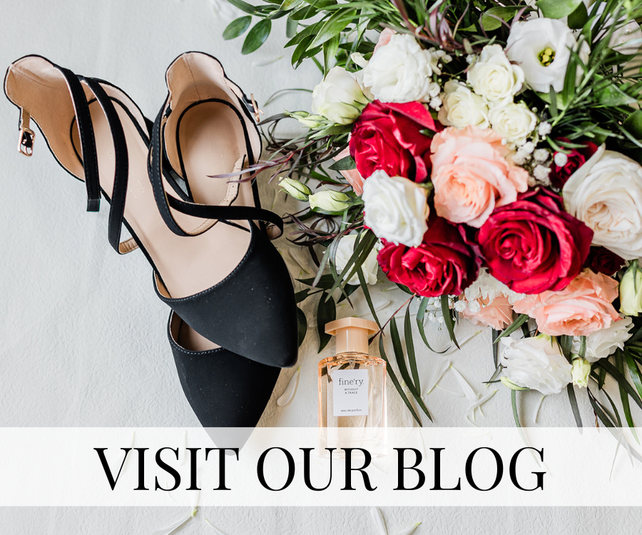 Black high-heeled shoes, a bottle of perfume, and a large bouquet of white, pink, and red roses with green foliage on a white surface. Text overlay says 'Visit Our Blog'.