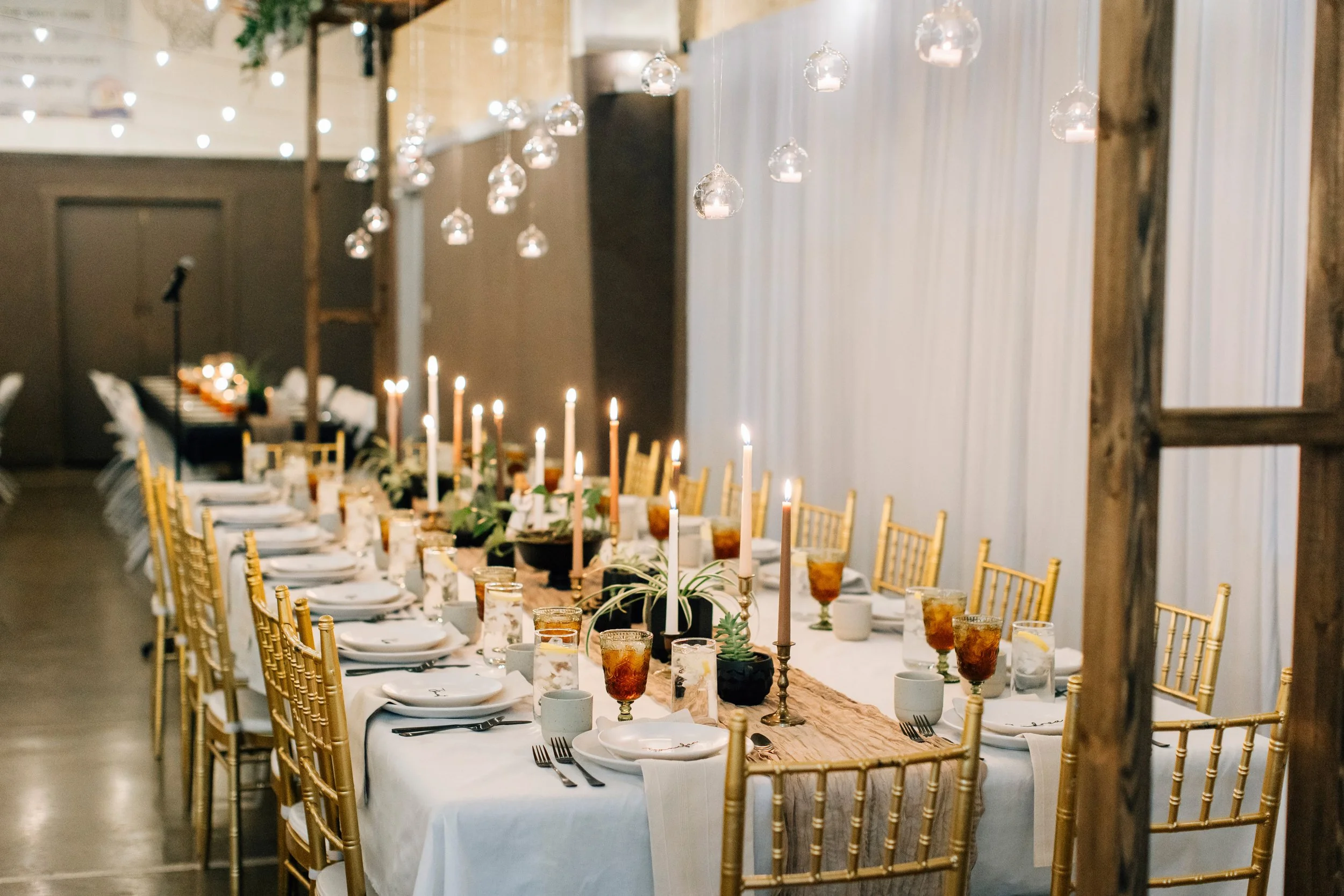 Elegant banquet table set with white tablecloth, gold chairs, candles, and glassware, decorated for a formal event in a rustic indoor venue with string lights and hanging glass ornaments.