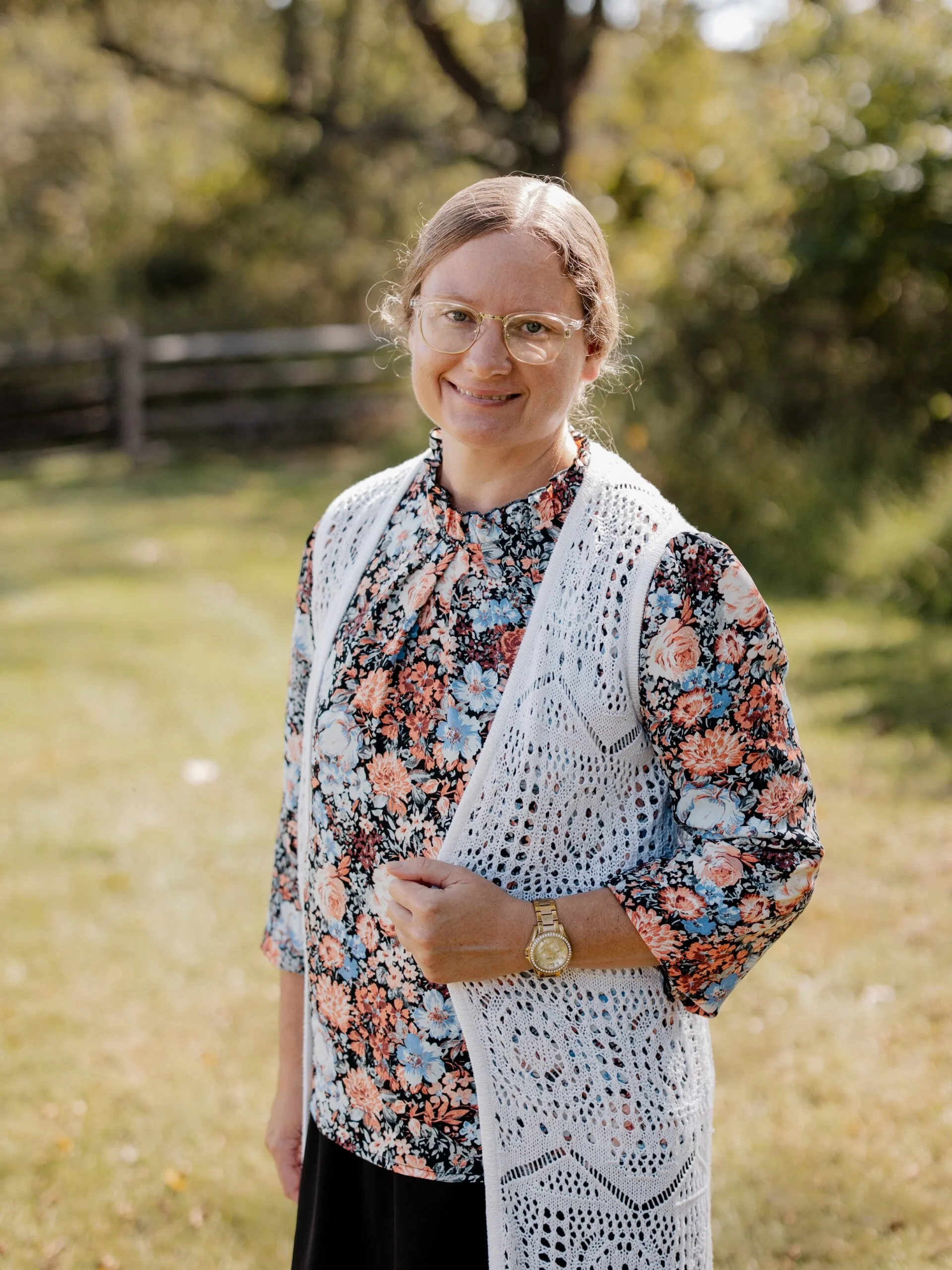 A woman with glasses wears a floral shirt and a white knitted vest, standing outdoors with trees and grass in the background.