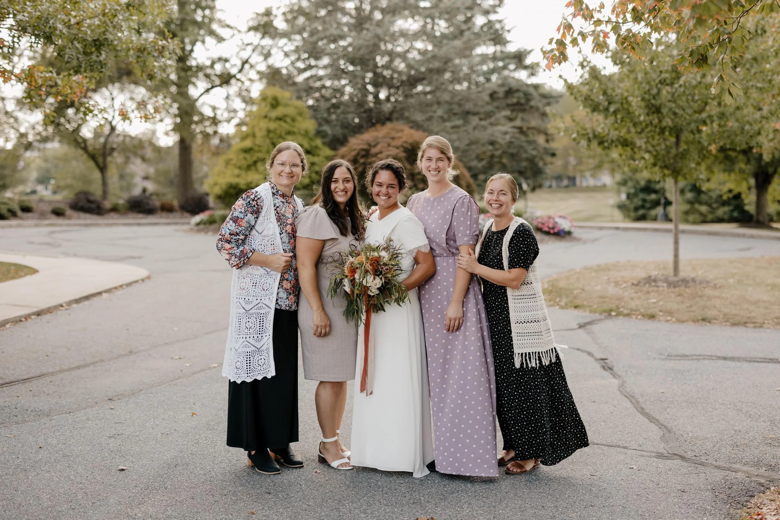 A group of five women standing outdoors in a park-like setting with trees and pathways, one woman in the center wearing a white dress holding a bouquet of flowers, smiling.
