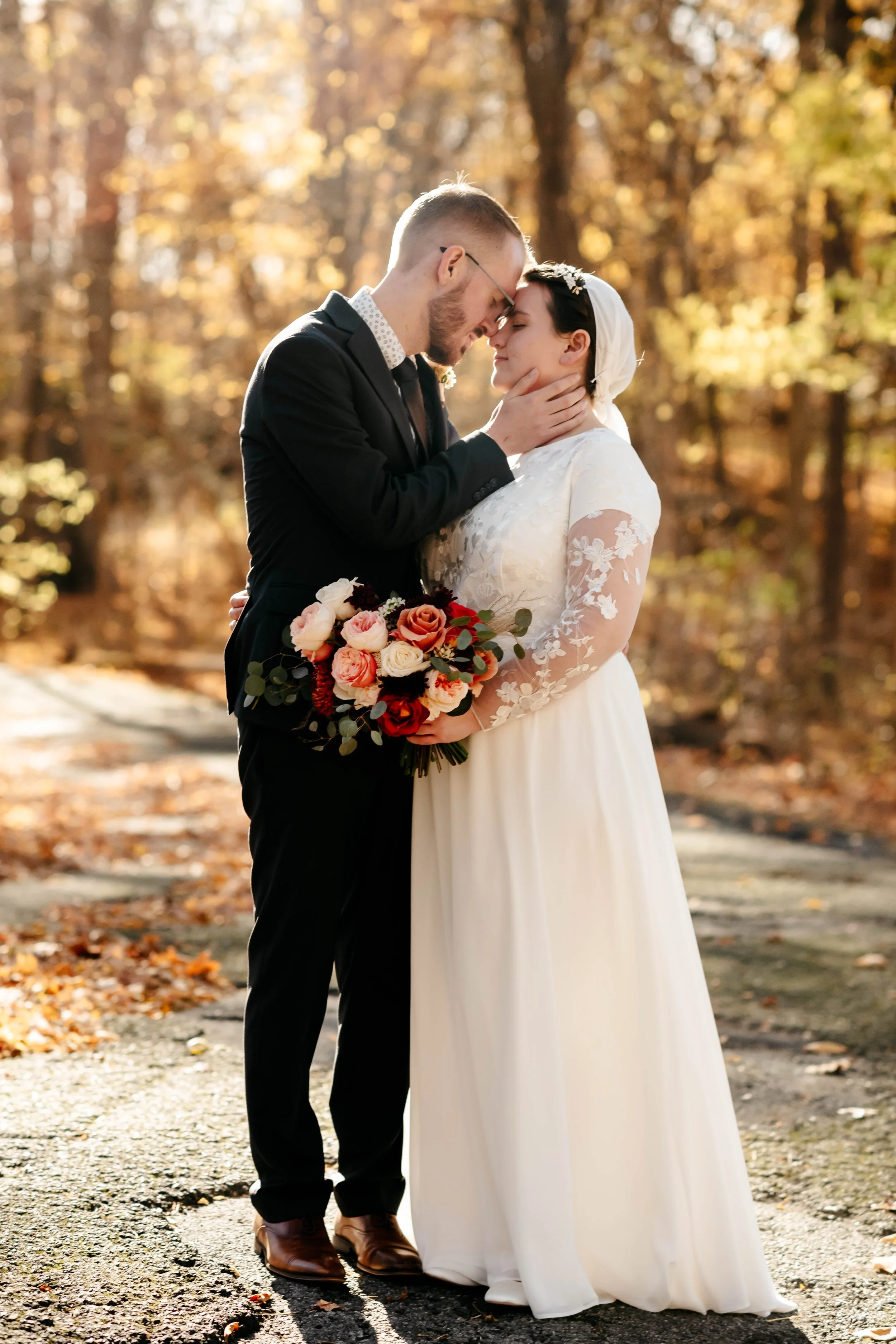 A newlywed couple sharing an intimate moment outdoors during autumn, with the groom gently holding the bride's face and the bride holding a bouquet of roses and greenery.