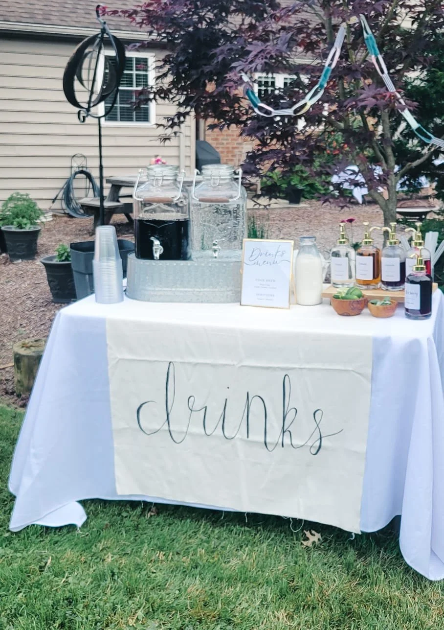 A drinks station set up outdoors in a backyard, featuring water dispensers, plastic cups, a milk jug, various flavored syrups, and a sign that reads 'drinks' on a table covered with a white tablecloth.