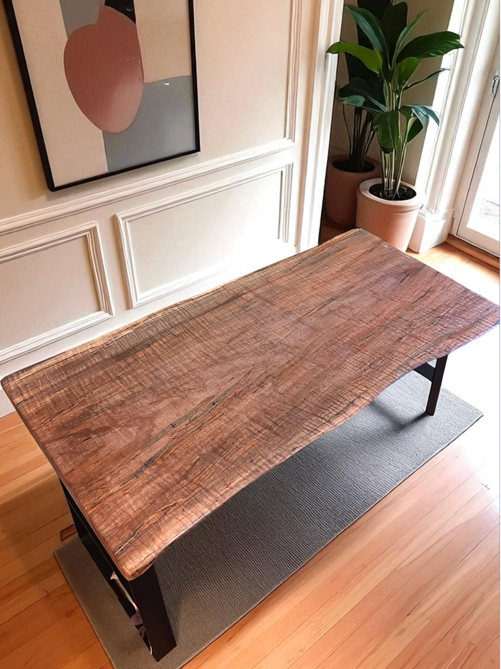 A wooden table with a natural, rustic finish in a room with beige walls, white wainscoting, and a framed abstract art piece. Two large potted green plants are on the windowsill near a glass door.