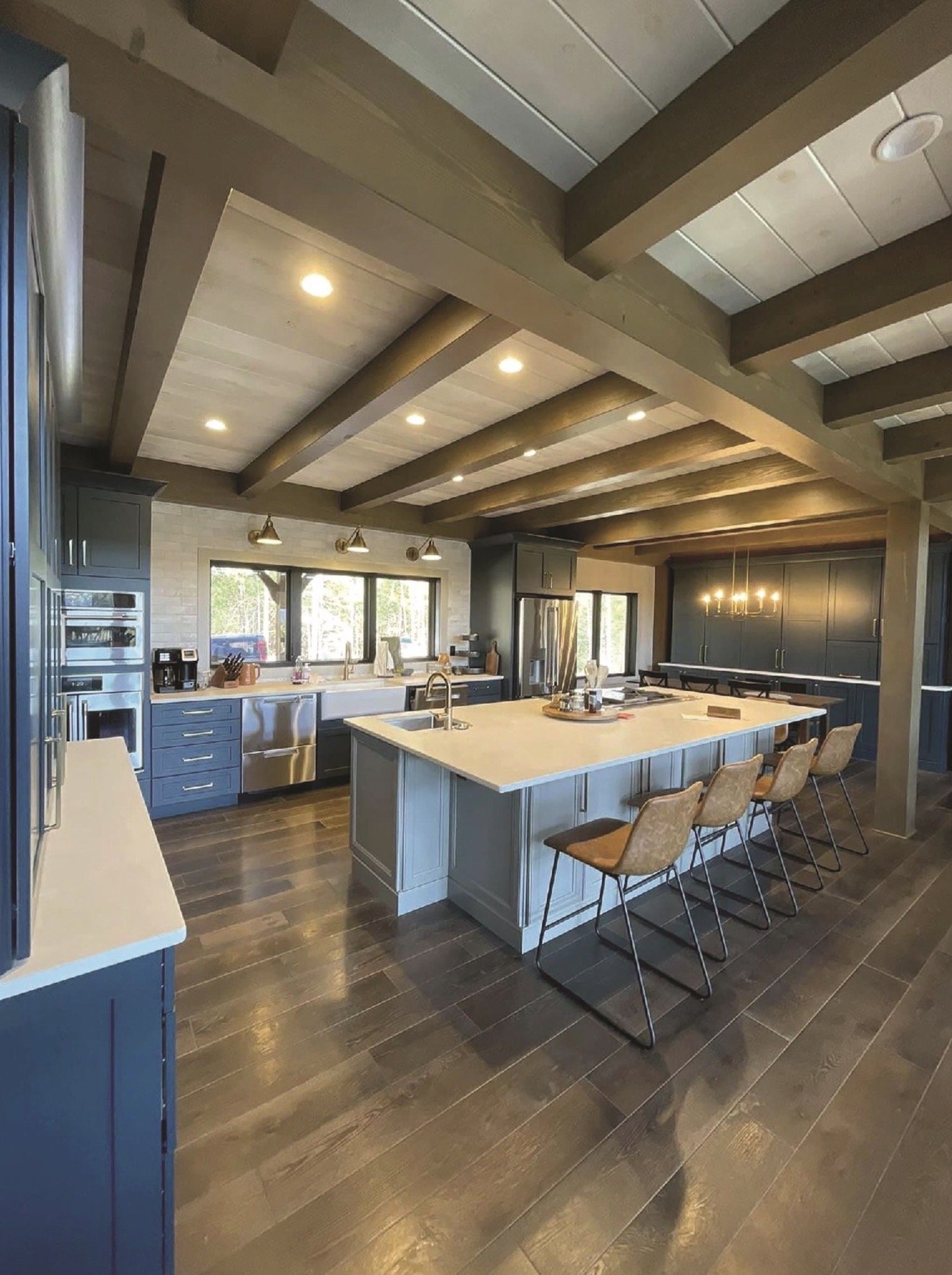 Modern kitchen with wooden ceiling beams, a large central island with four chairs, dark blue cabinets, stainless steel appliances, and a chandelier over the dining area.