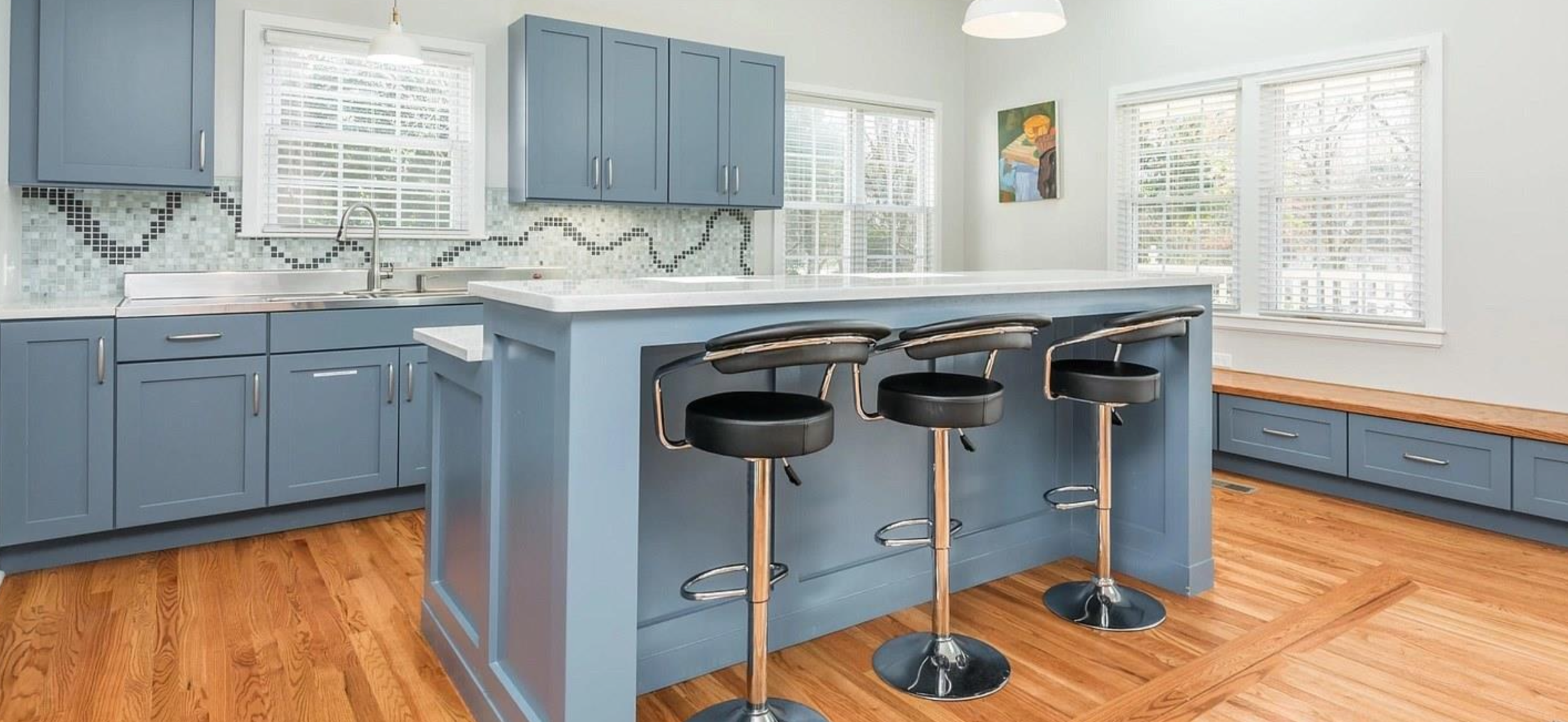 Bright kitchen with blue cabinets, a white countertop, a tile backsplash, large windows with blinds, a wooden bench, and three black stools at a kitchen island.