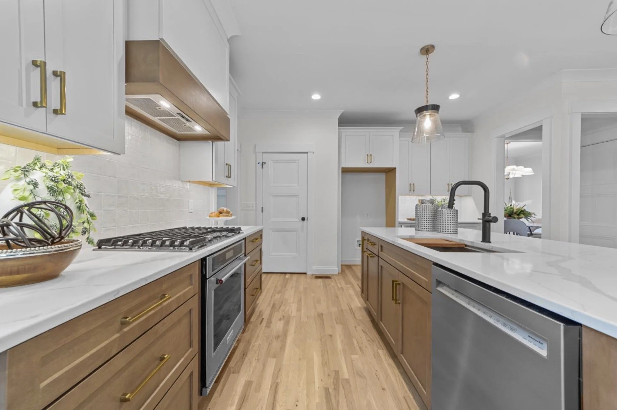 Modern kitchen with white upper cabinets, wooden lower cabinets, stainless steel appliances, a white marble countertop island with a black faucet, hanging pendant light, and light wooden flooring.