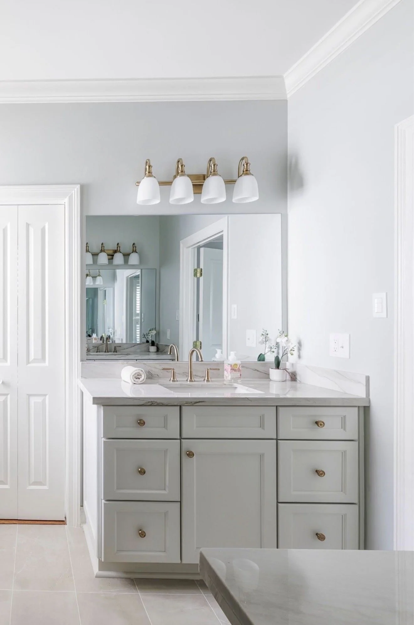 Bathroom vanity with a marble countertop, a large mirror, and a three-bulb brass light fixture. Light gray cabinetry and decor accentuate a clean, modern aesthetic.