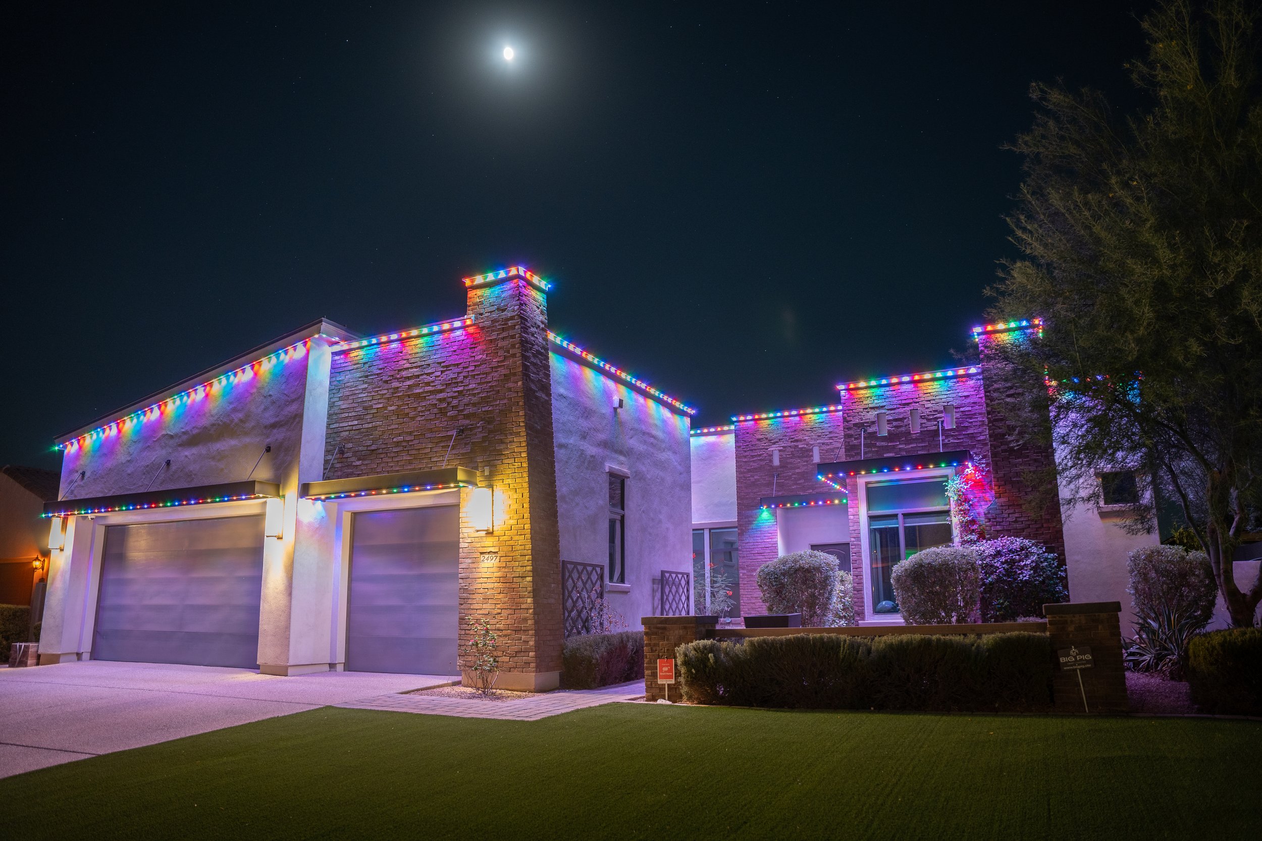 A modern house decorated with colorful Christmas lights at night, with a bright moon in the night sky.