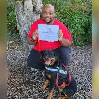A man sitting outdoors next to a service dog, holding a certificate.