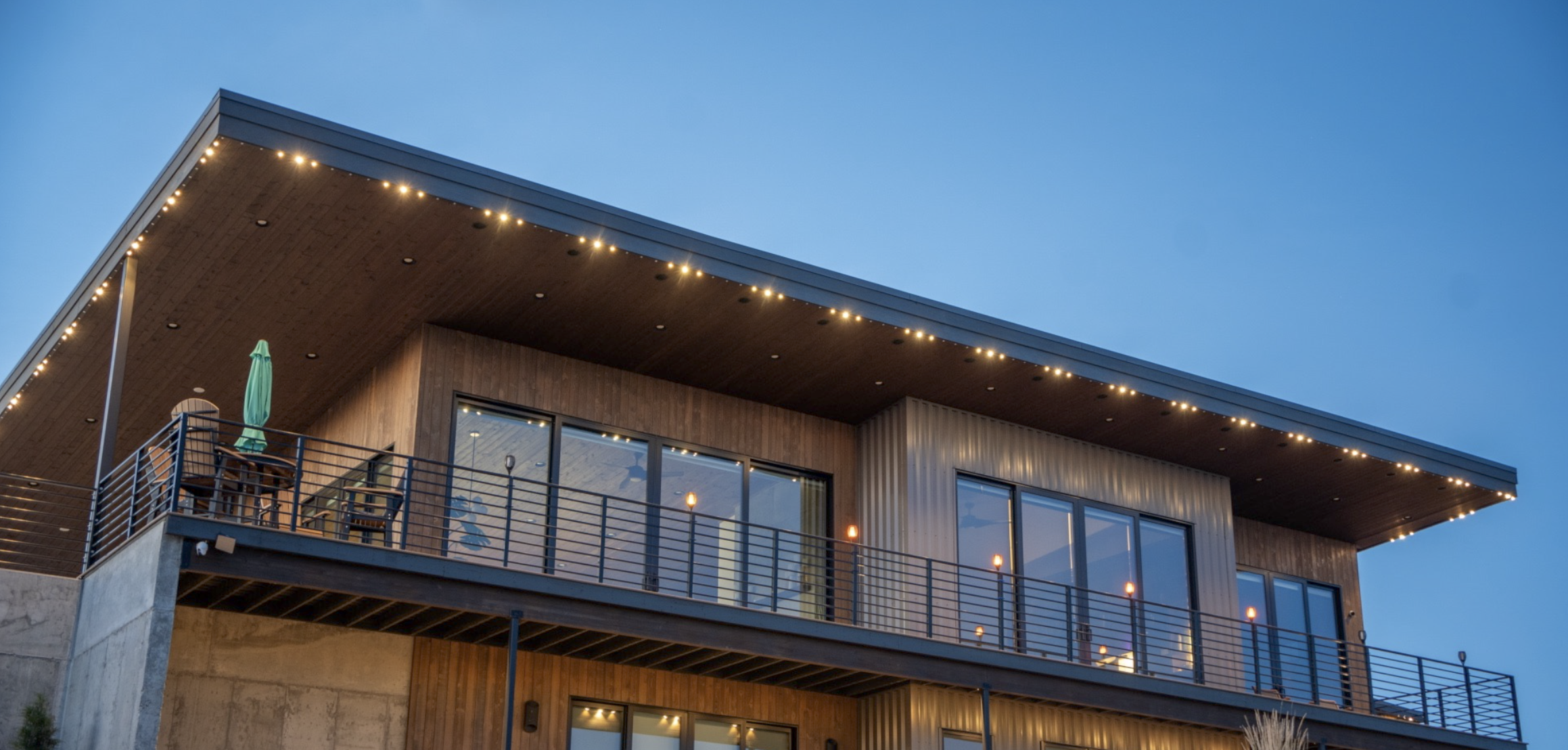 Modern house with large glass windows and a balcony, illuminated with string lights, under a clear blue evening sky.