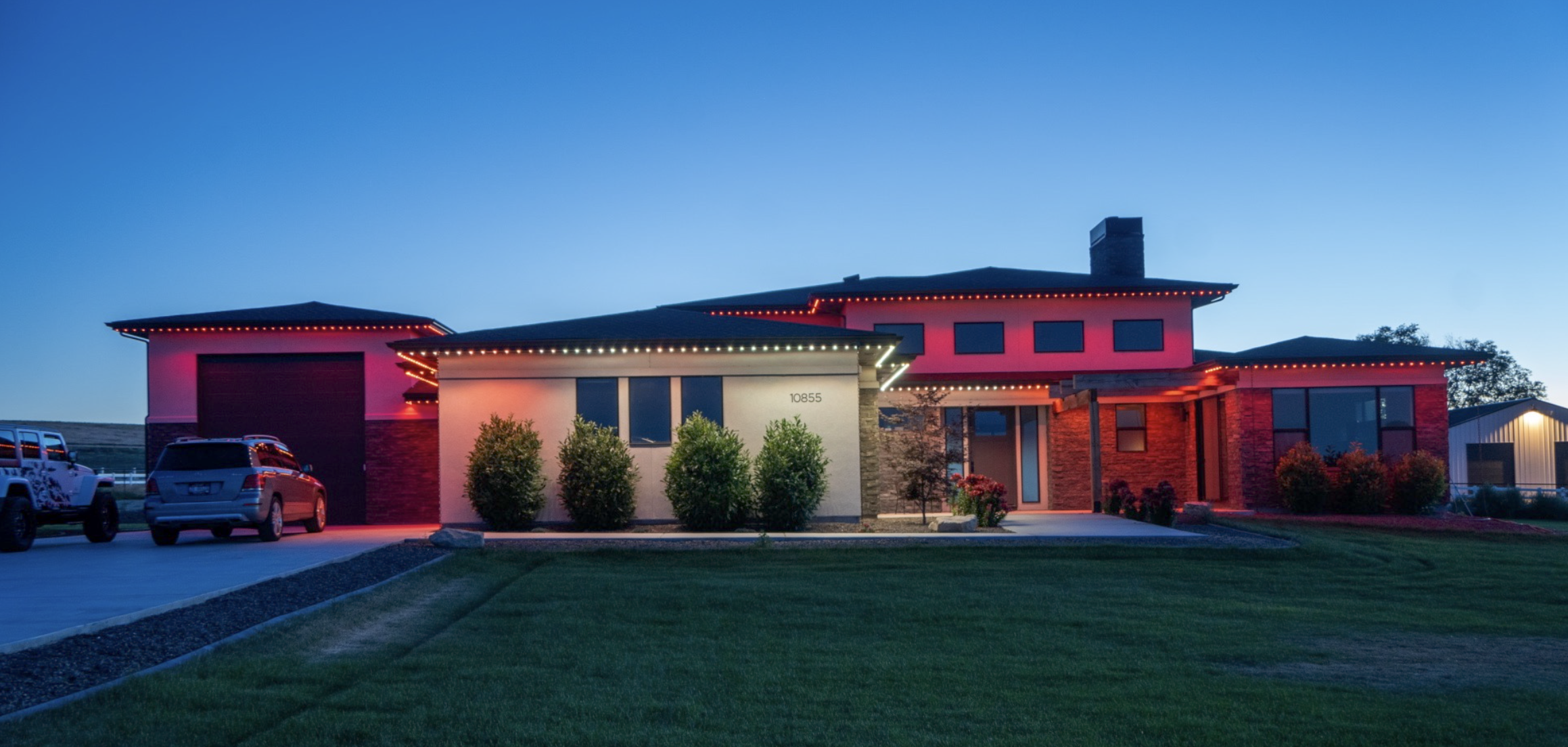 Modern house with red and white exterior, decorated with red and white string lights, parked cars nearby, manicured lawn in front, during twilight.