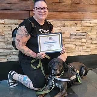 Person kneeling with a black dog in front of a stone wall, holding a framed certificate, wearing glasses and a black T-shirt.