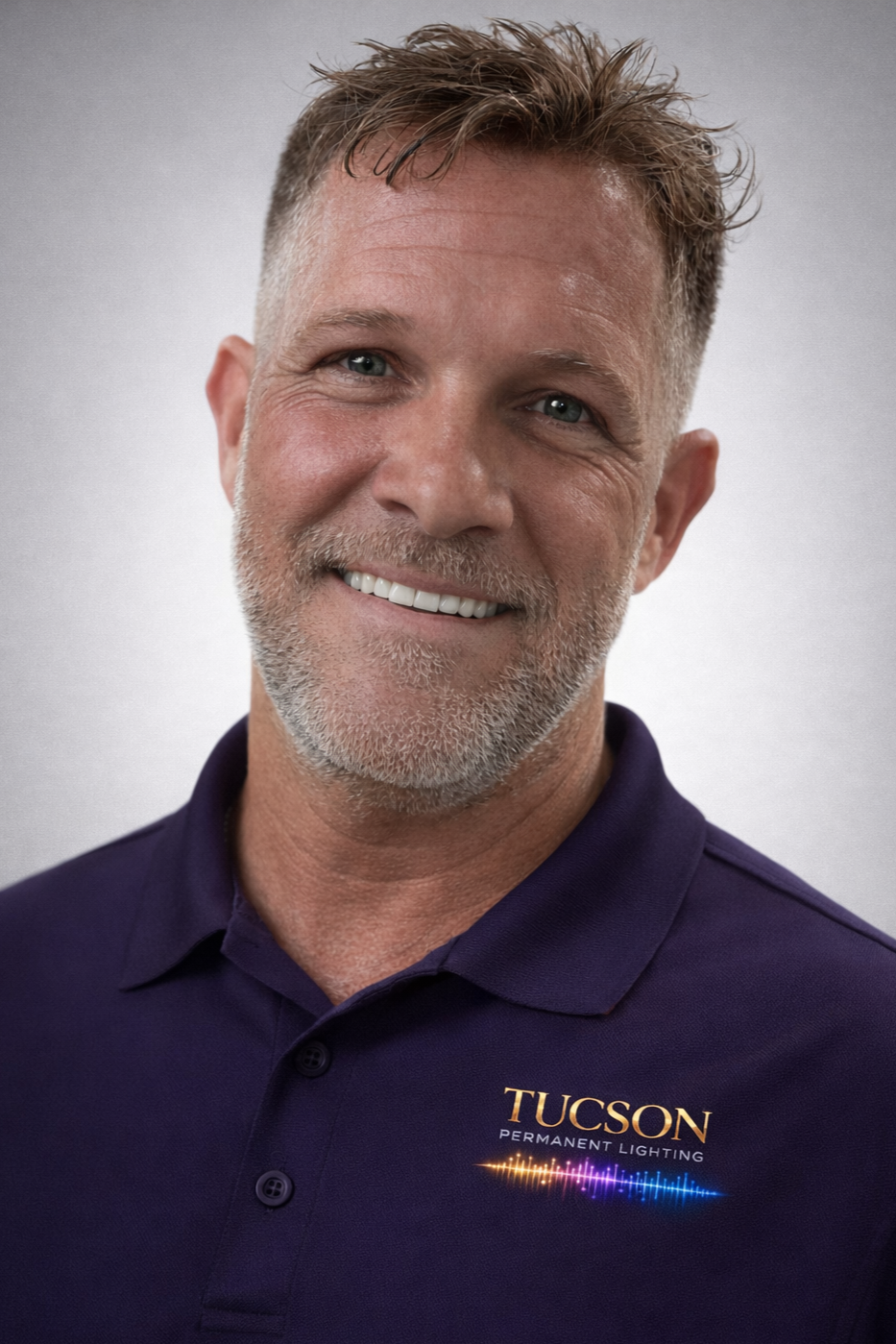 A middle-aged man with short gray hair and a beard, smiling, wearing a dark purple polo shirt with the logo 'TUCSON PERMANENT LIGHTING' on it.