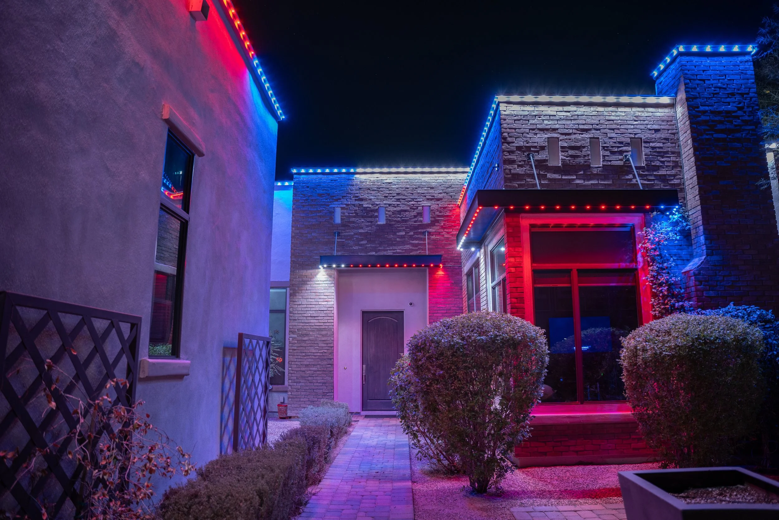 Night view of a modern house with exterior colorful neon lights illuminating the walls and roofline, surrounded by neatly trimmed bushes and a brick pathway.