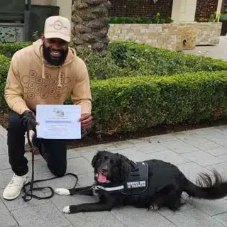 A man smiling and holding a certificate beside a black service dog lying on the sidewalk outdoors.