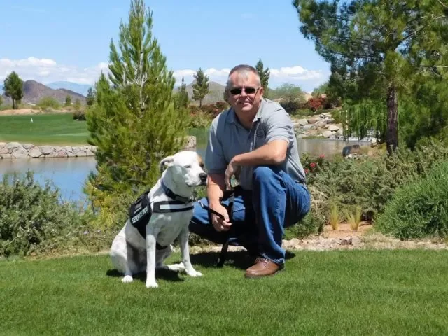 Man crouching next to a white service dog in a park with trees, grass, and a body of water in the background.